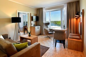 A modern hotel room with a wooden floor, a sofa, a dining table, a TV, and a view of a lake.