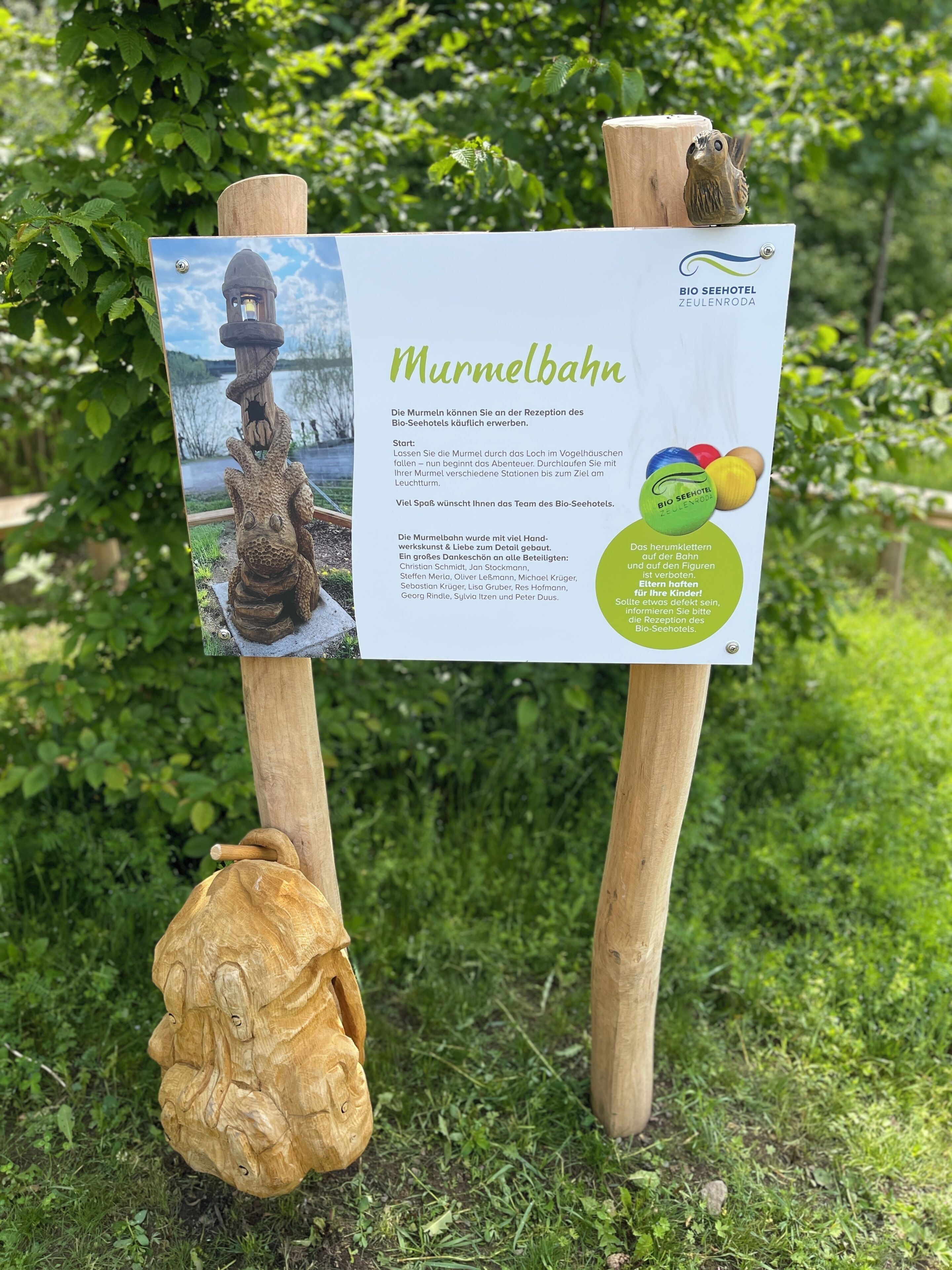 Un letrero de madera con texto y una fotografía de una escultura de madera, sobre un fondo de vegetación.
