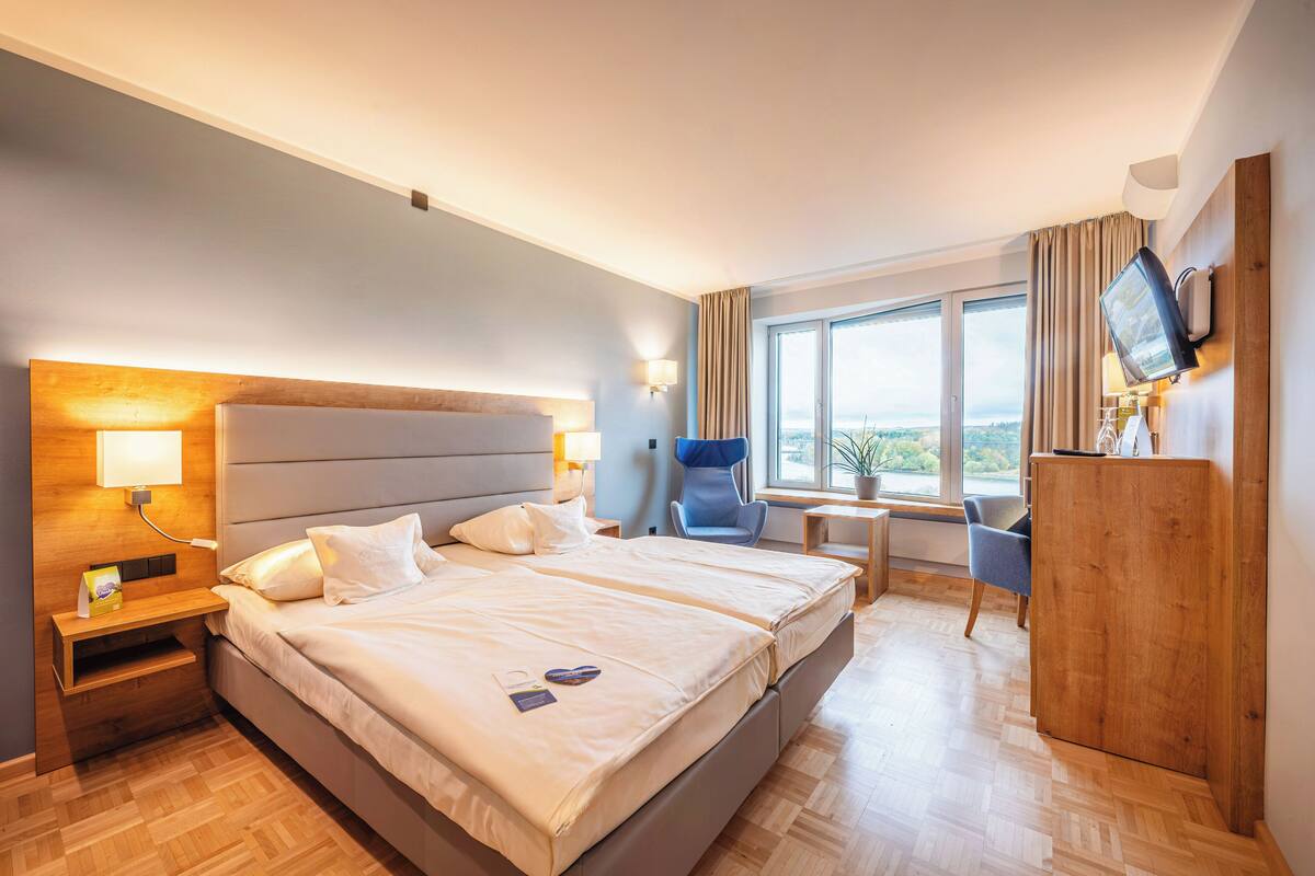 A hotel room with a large bed, bedside tables, a desk, a chair, a TV, and a window with a view.