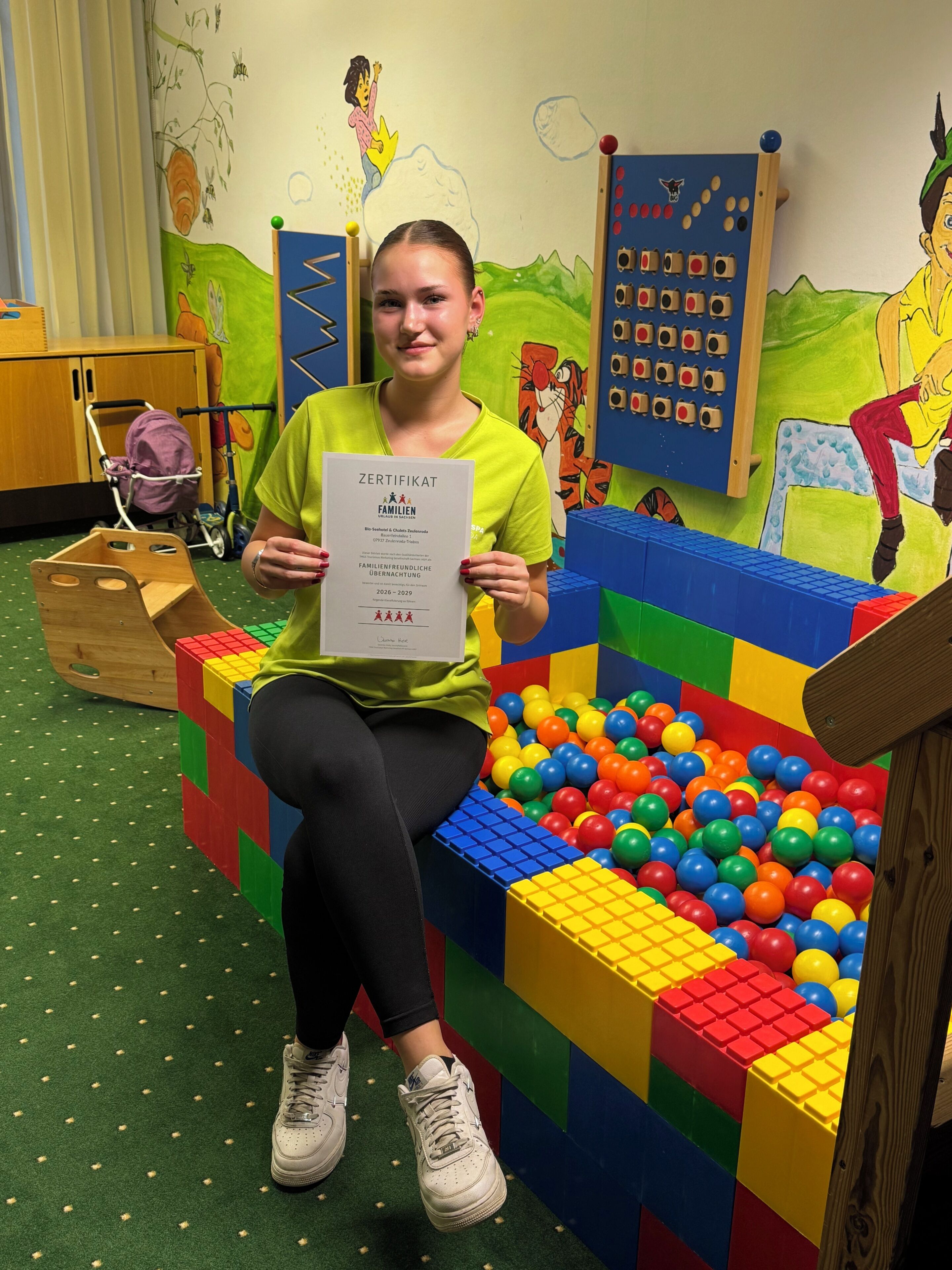 A child sitting on a colorful play structure in a room with a green carpet, wooden furniture, and educational wall decorations.