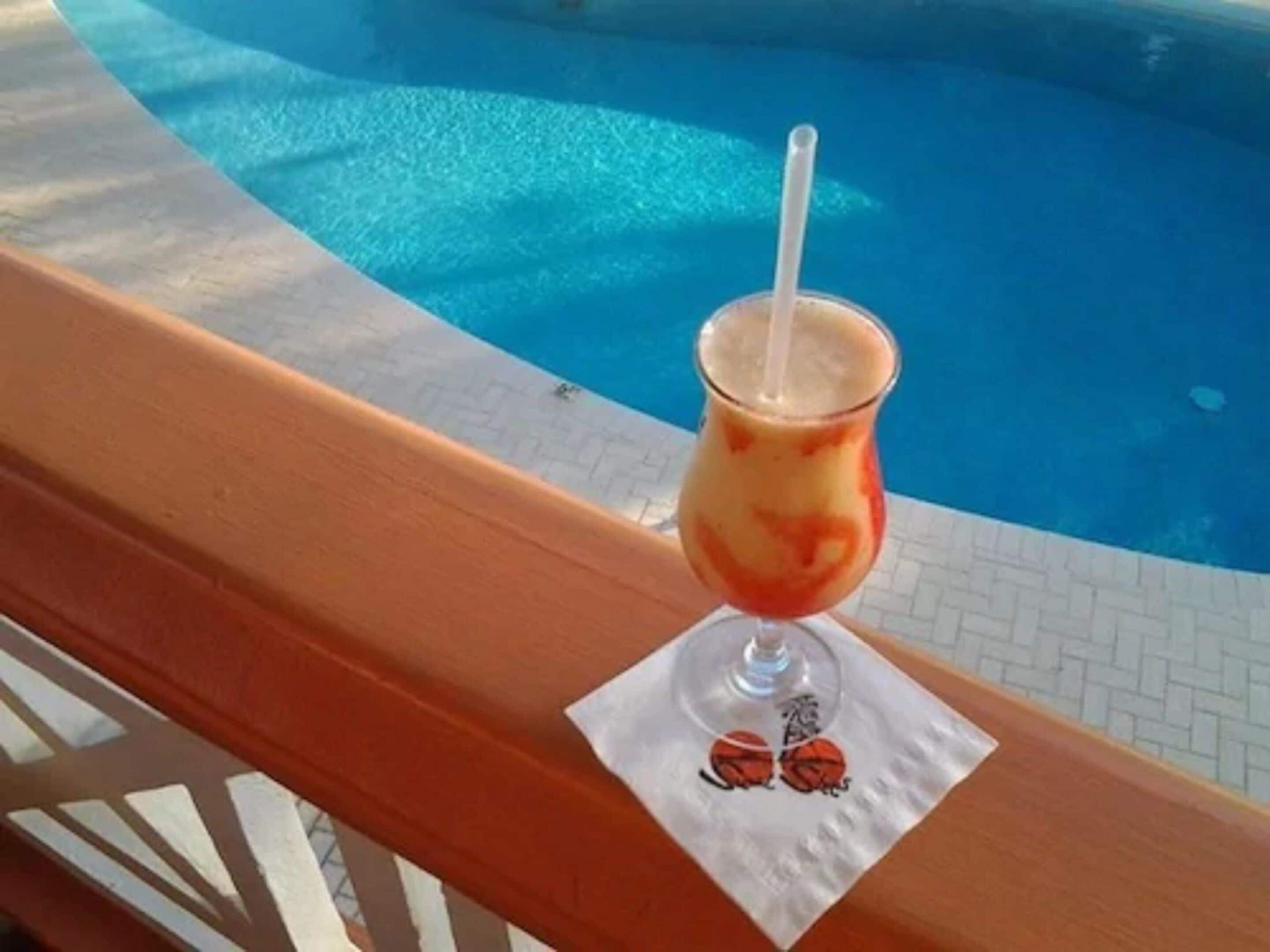 Ein Glas mit einem roten Getränk und einem Strohhalm auf einer Holzterrasse an einem Pool.
