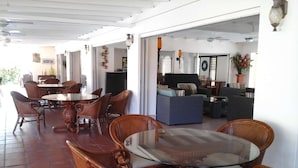 Uma ampla área de refeições ao ar livre com cadeiras de vime e uma mesa com tampo de vidro, que dá acesso a uma sala de estar interna com sofás e uma televisão.