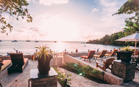 A beachfront patio with lounge chairs, a potted plant, and a view of the sea with boats.