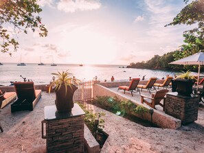 A beachfront patio with lounge chairs, a potted plant, and a view of the sea with boats. - Sunset Shores Beach Hotel (Villa)