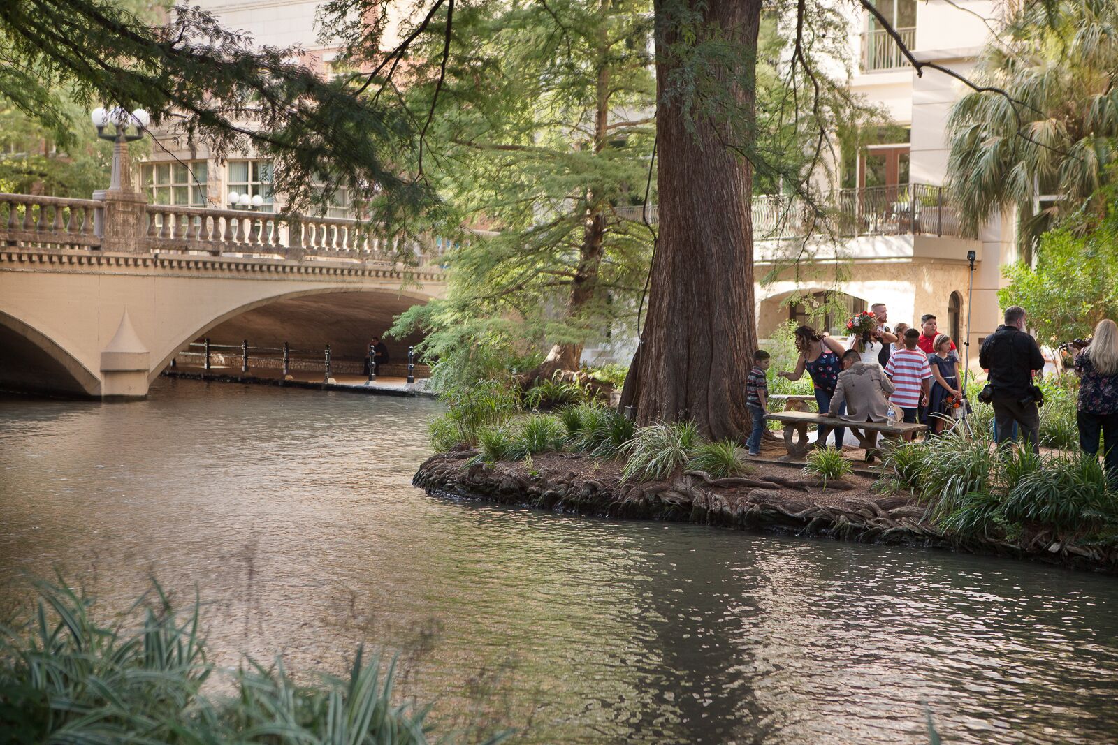A riverside area with a stone bridge, lush greenery, and a group of people gathered around a table.