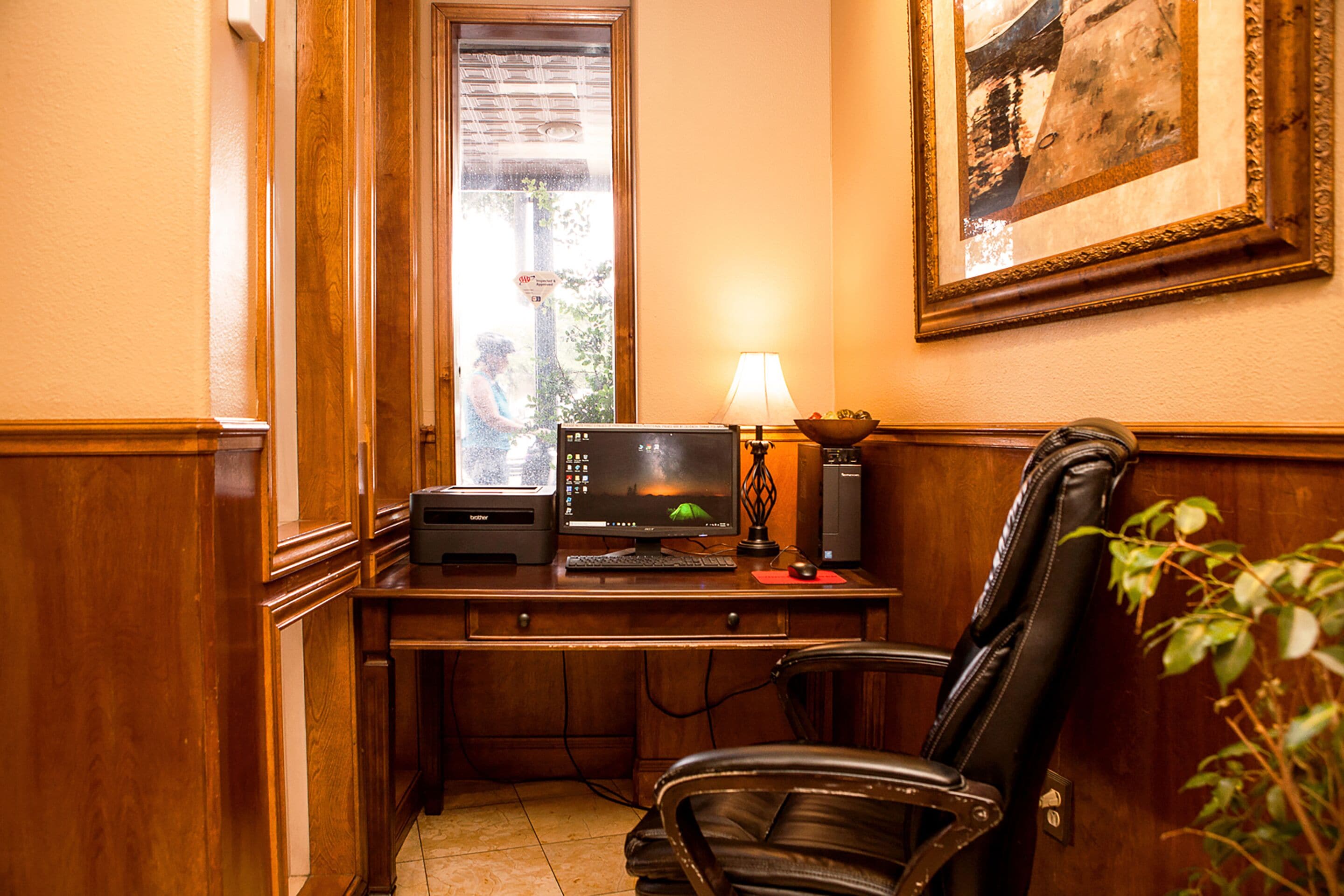 Un bureau en bois avec un ordinateur, une lampe et une chaise, dans une pièce dotée d’une porte et d’un tableau encadré au mur.