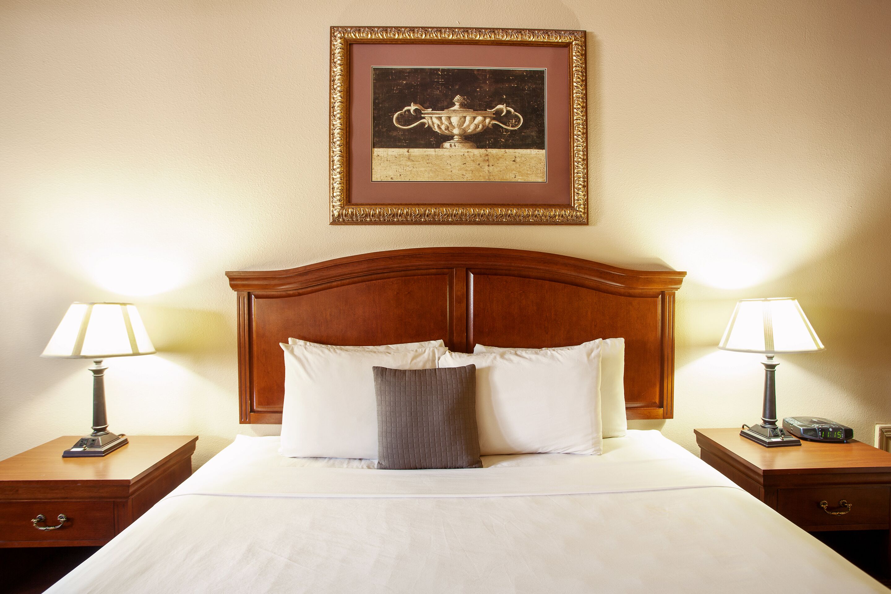 A neatly made bed with white linens, two bedside tables with lamps, a framed picture above the bed, and a wooden headboard.