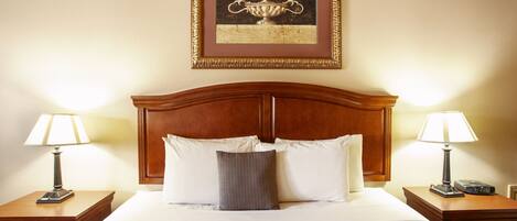 A neatly made bed with white linens, two bedside tables with lamps, a framed picture above the bed, and a wooden headboard.