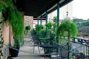 Un patio al aire libre con mesas y sillas de metal, rodeado de plantas en macetas y una valla de malla.