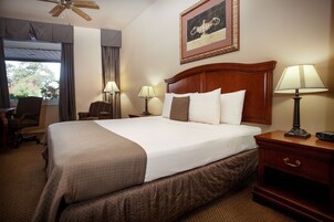 Ein Hotelzimmer mit einem großen Bett, zwei Nachttischlampen, einem Holznachttisch, einem Sessel und einem Fenster mit Vorhängen.
