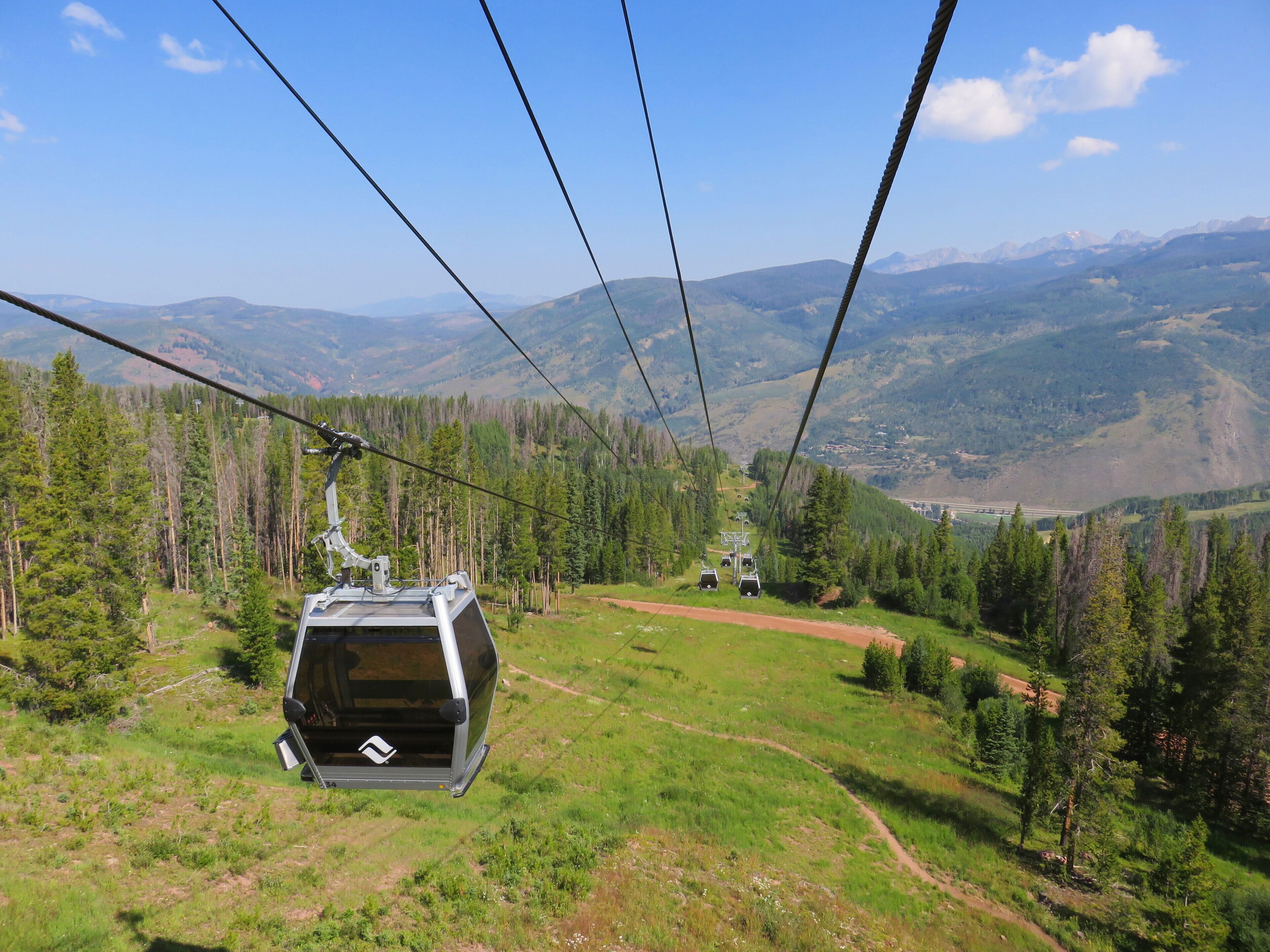 A cable car moving over a mountainous landscape with green fields and forested areas.