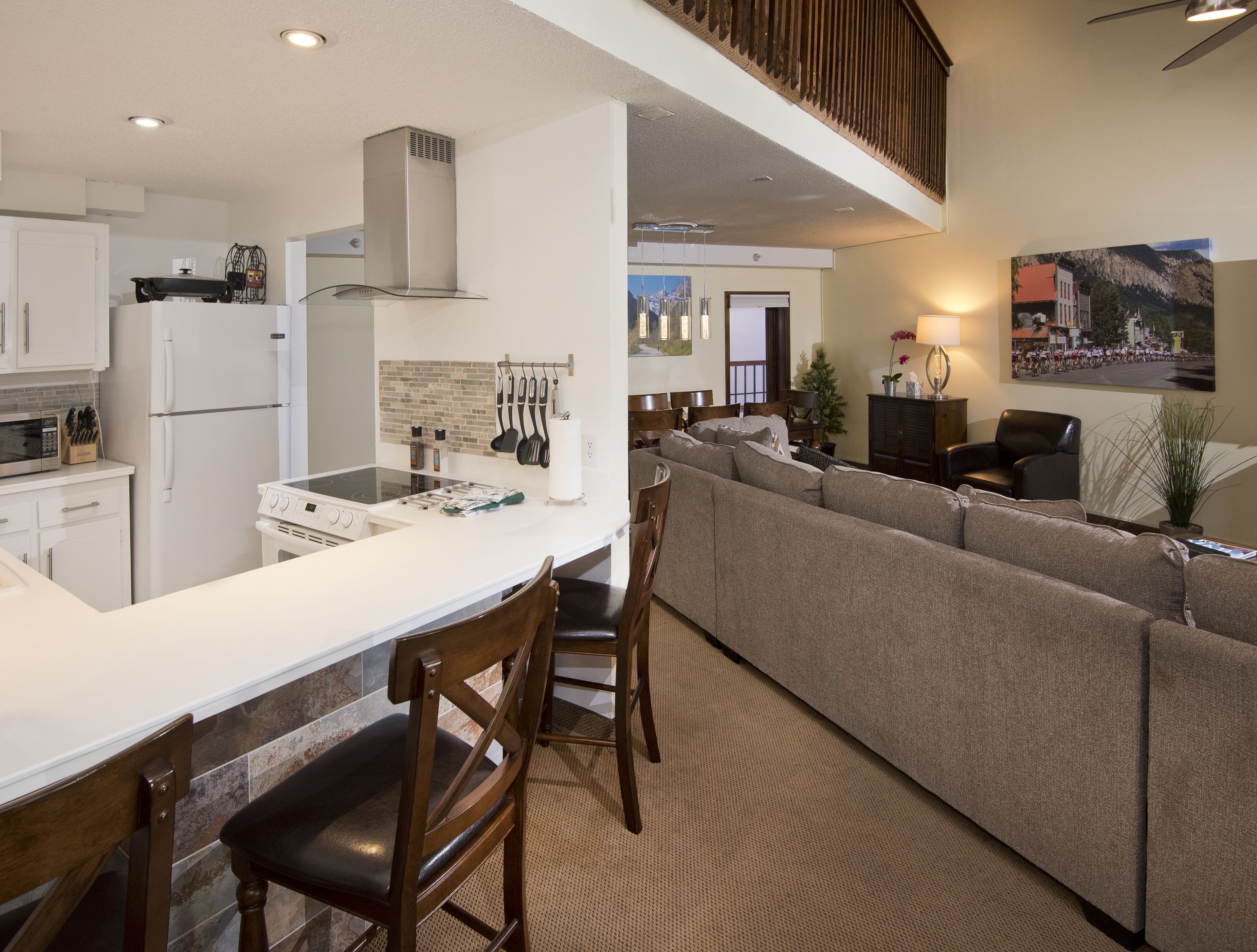 A modern kitchen with white appliances, a dining area with a white table and chairs, and a living area with a large sofa and a TV.