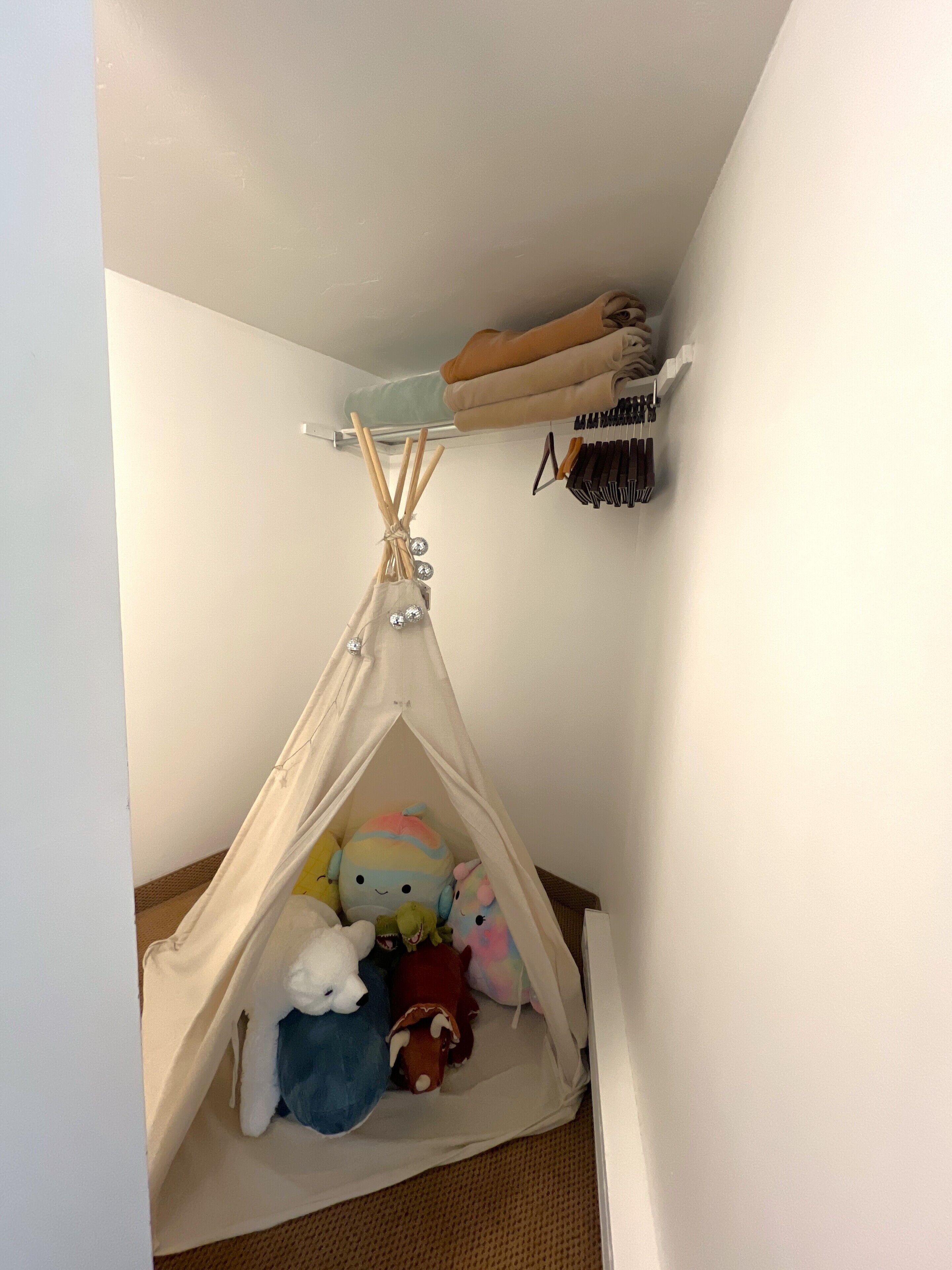 A corner room with a white wall, a beige tent-like structure with stuffed toys, a shelf with folded clothes, and a hook with black items.