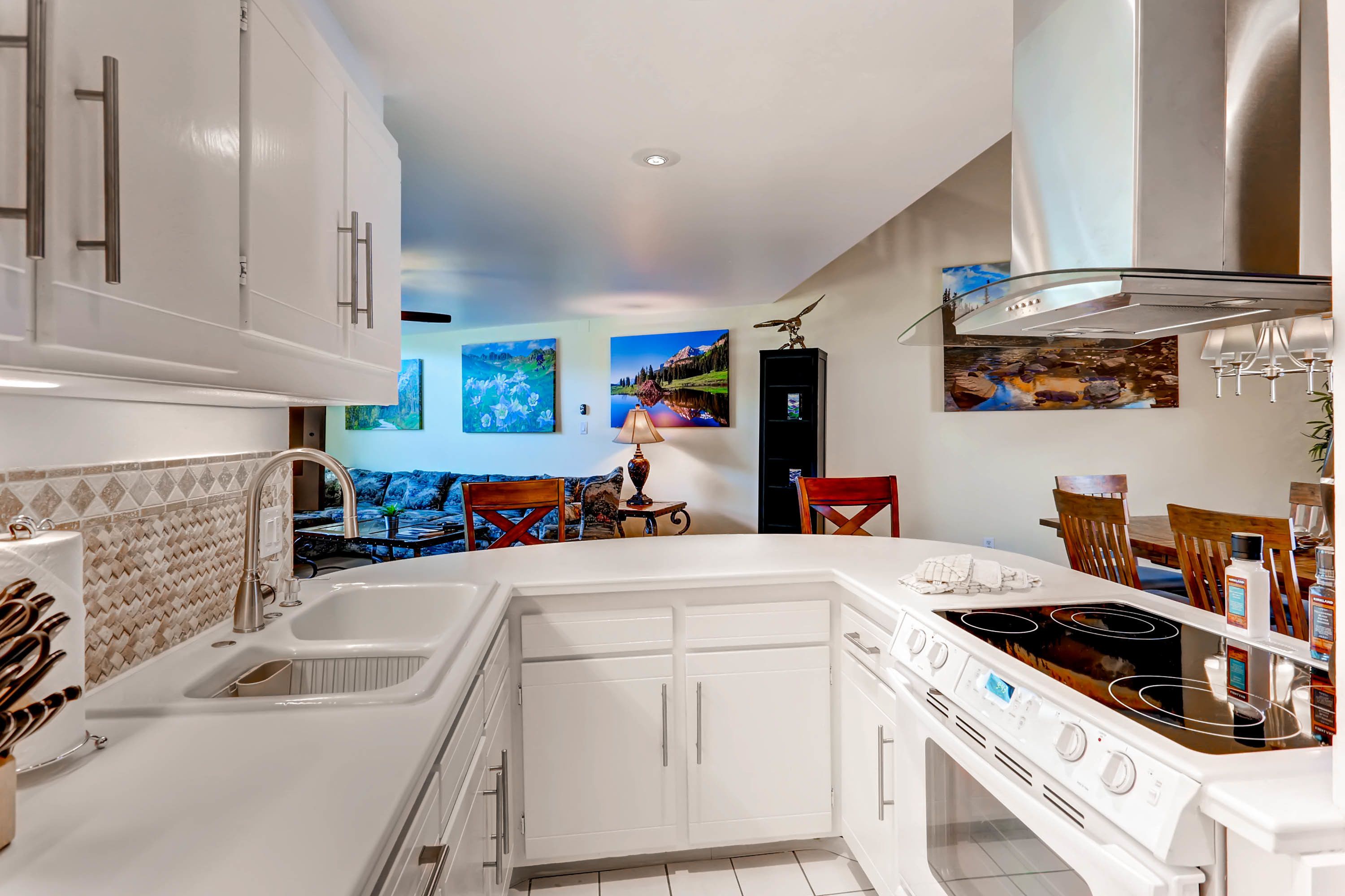 A modern kitchen with white cabinetry, a double sink, and a gas stove. There are decorative paintings on the walls and a dining area with wooden chairs.