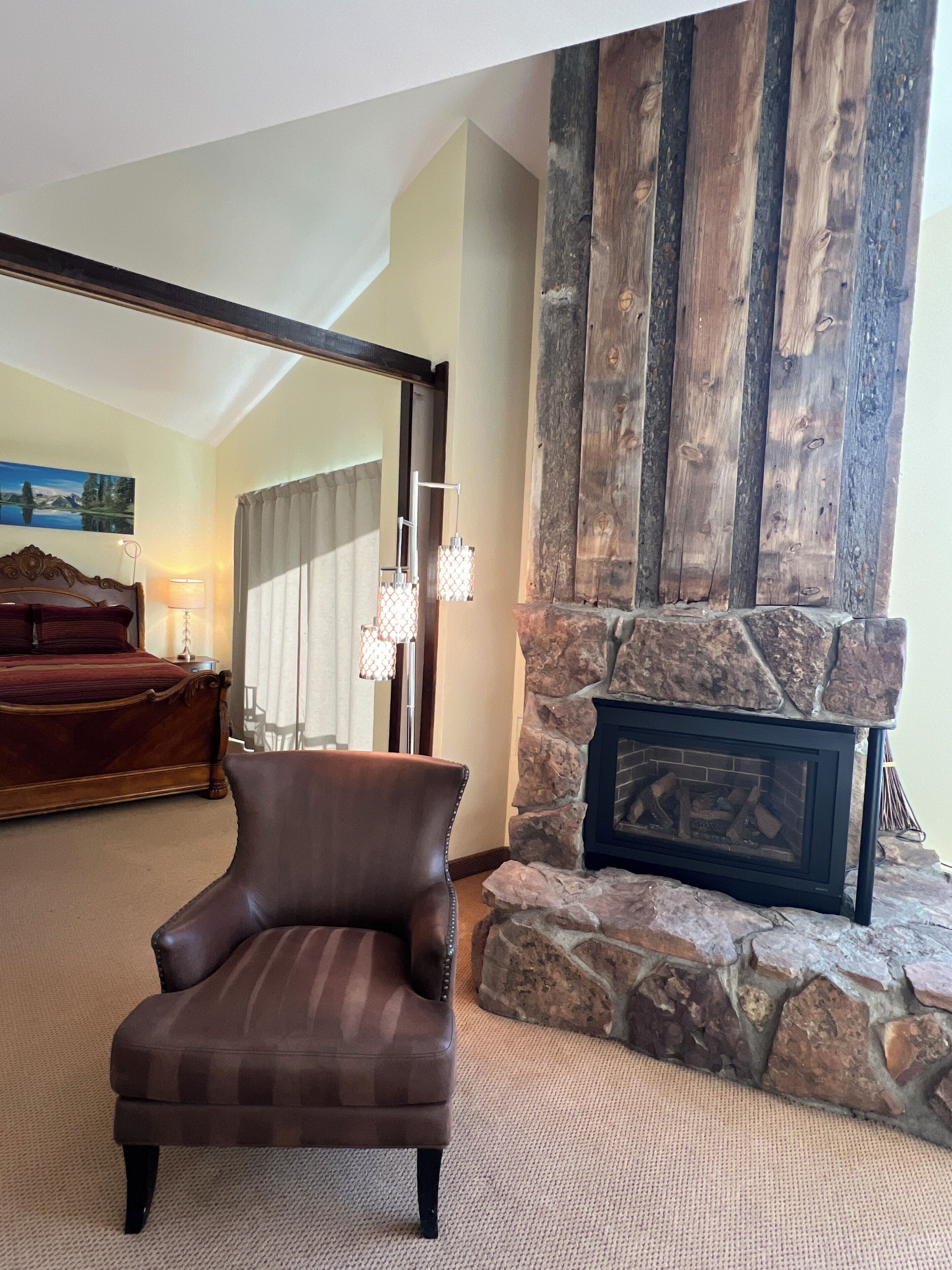 A room with a stone fireplace, a leather armchair, a bed with a wooden headboard, and a painting on the wall.