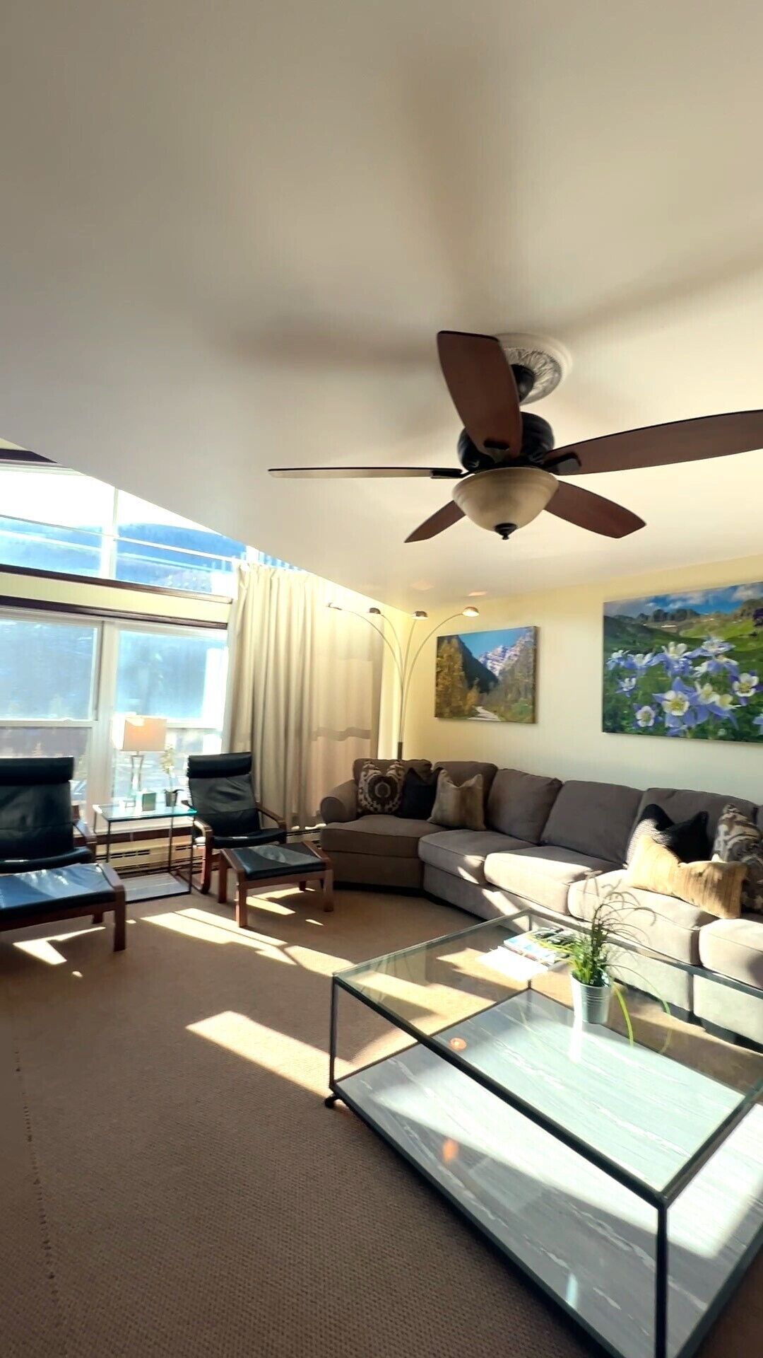 En moderne stue med ventilator, sofa, sofabord i glas og et maleri på væggen.