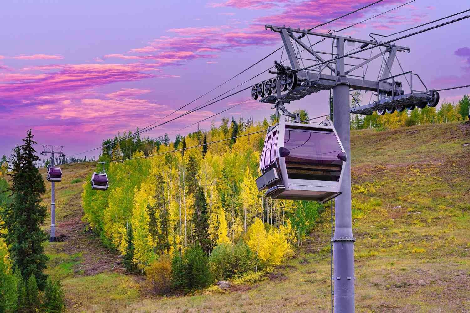 Eine Seilbahn mit Gondeln, die an einer Metallkonstruktion befestigt sind, umgeben von einem Wald mit herbstlich gefärbten Bäumen.