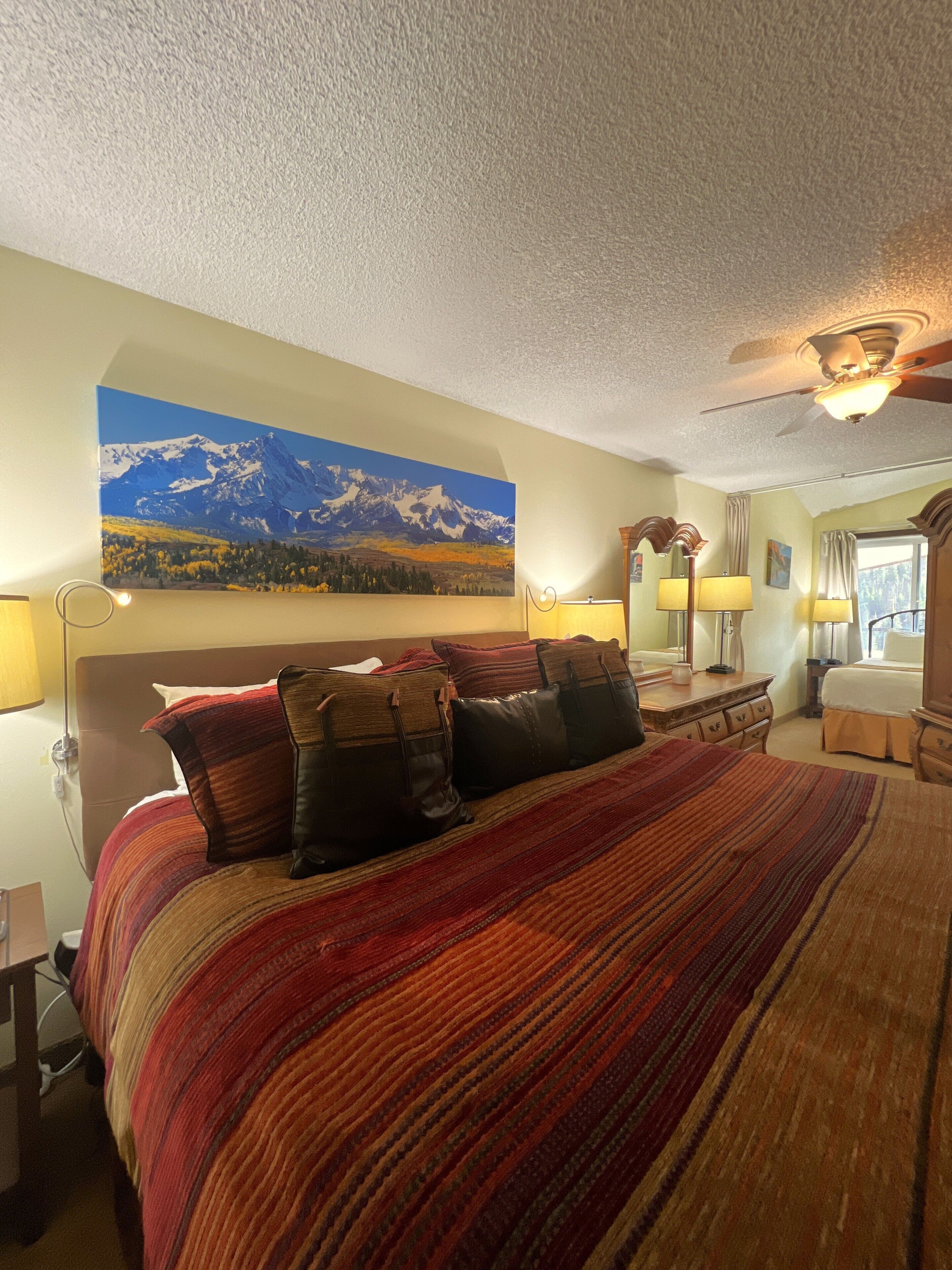 Une chambre d’hôtel avec un grand lit, un tableau représentant des montagnes, un ventilateur de plafond et une fenêtre avec des rideaux.