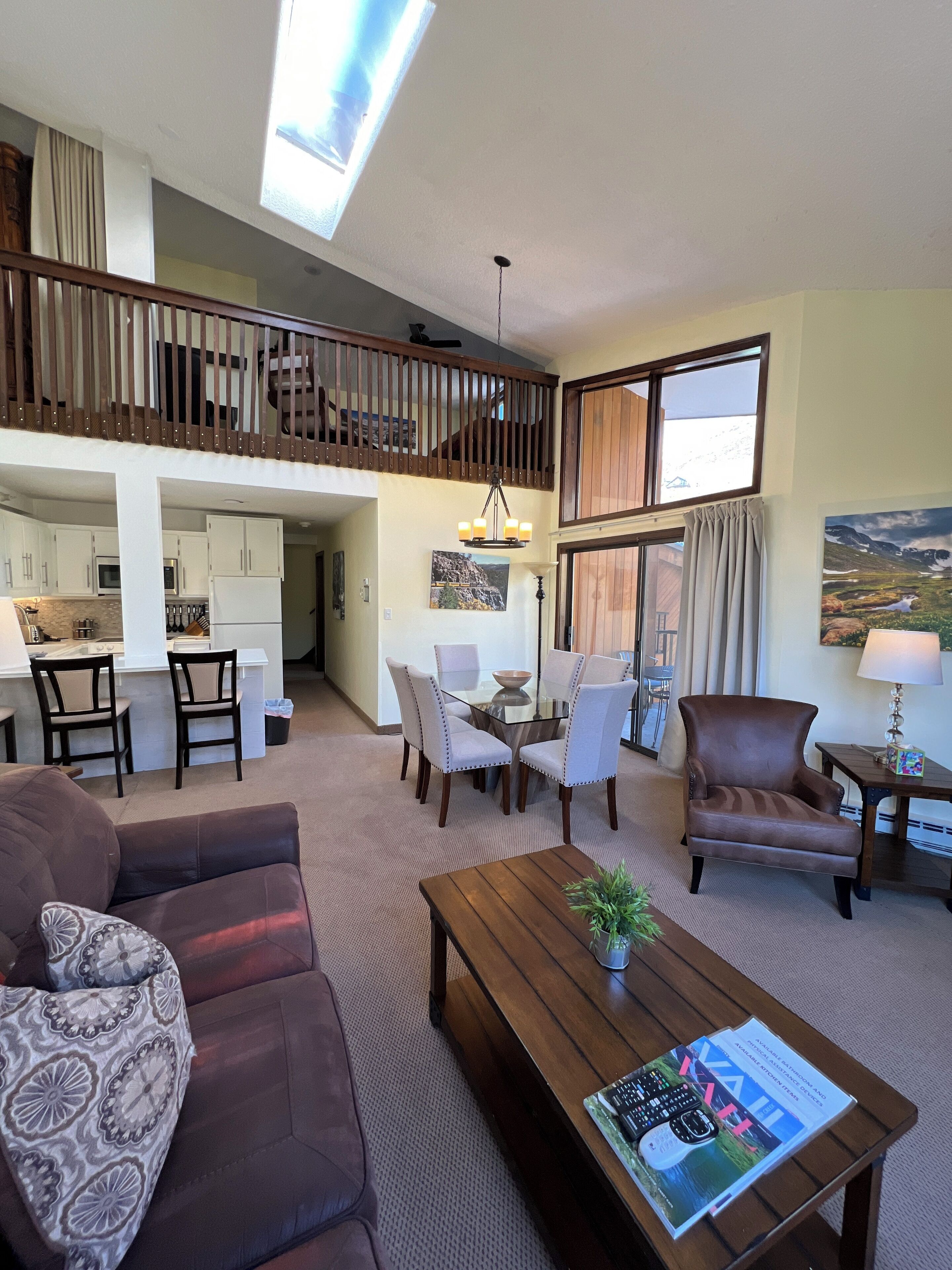 A spacious living area with a high ceiling, a dining table with chairs, a brown leather sofa, a wooden coffee table, and a view of the kitchen and balcony.
