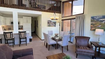 A spacious living area with a high ceiling, a dining table with chairs, a brown leather sofa, a wooden coffee table, and a view of the kitchen and balcony.