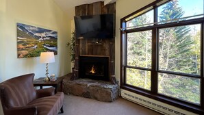 A cozy living room with a fireplace, a television mounted above it, and a large window offering a view of trees. - Vail Run Resort (Vail)