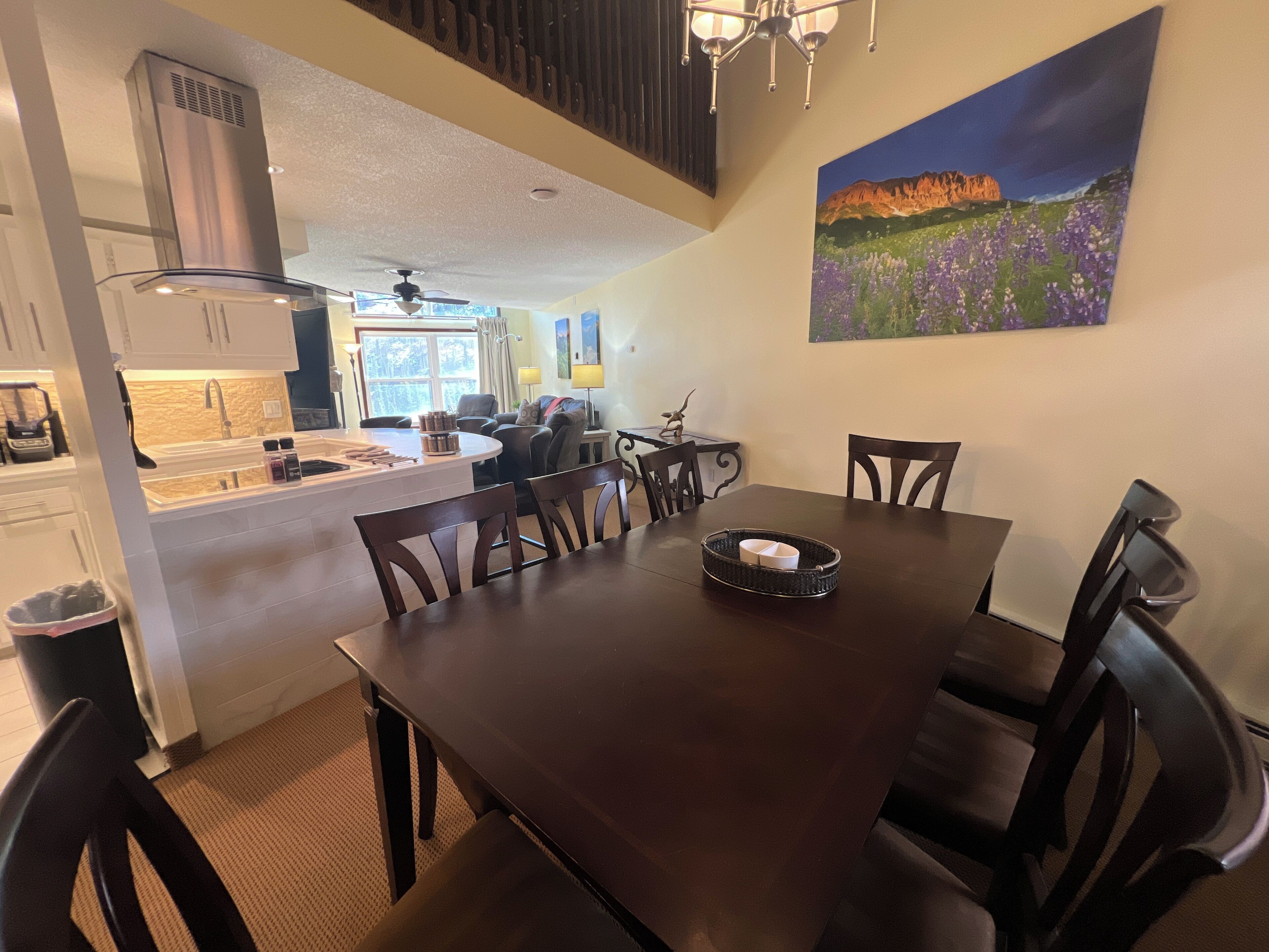 A dining area with a dark wooden table and chairs, a kitchen in the background, and a painting of a mountain landscape on the wall.