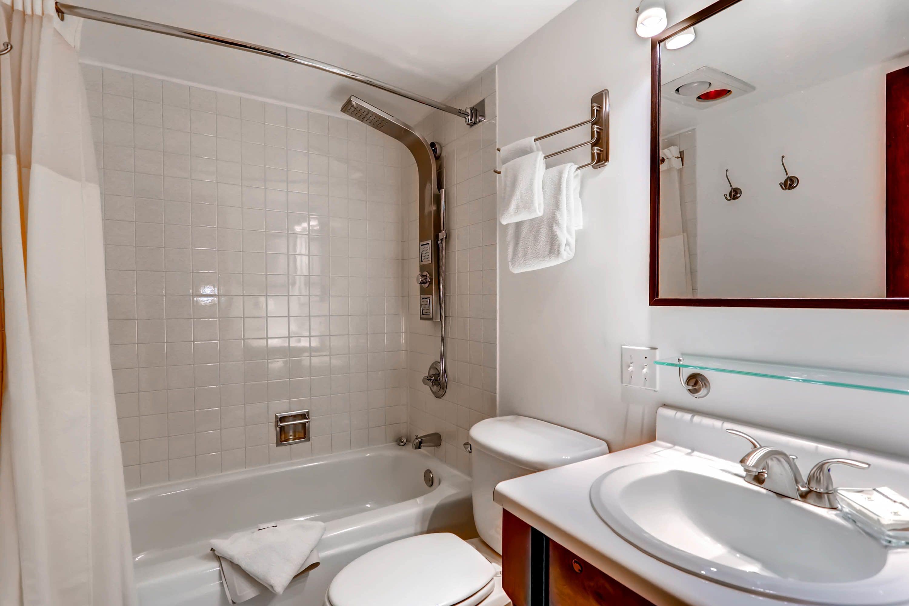 Une salle de bain moderne équipée d’une baignoire, de toilettes et d’un lavabo, avec des carreaux blancs et une étagère en verre.