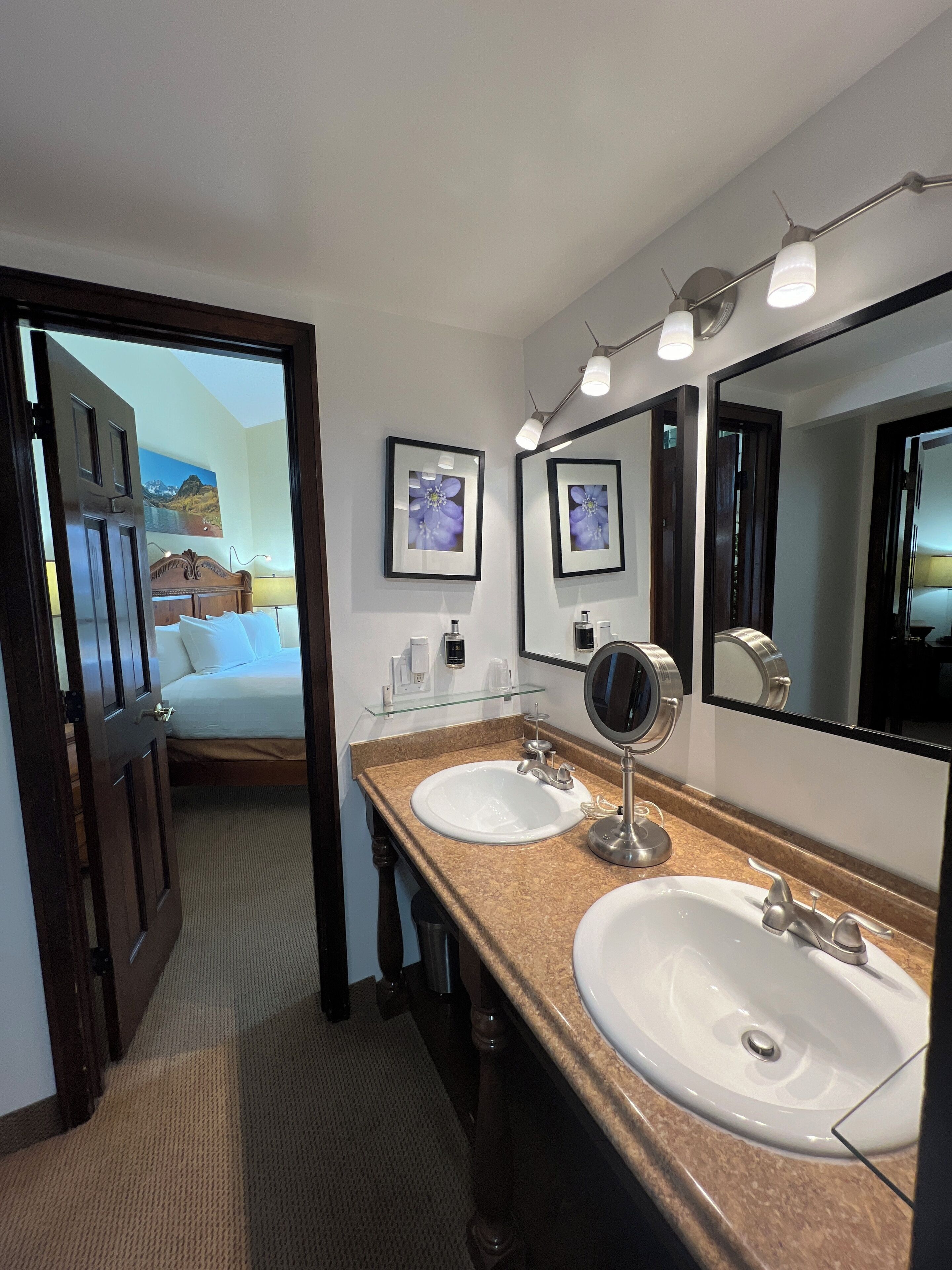 Uma casa de banho de hotel com duas pias, um espelho grande e uma cama visível através de uma porta aberta.