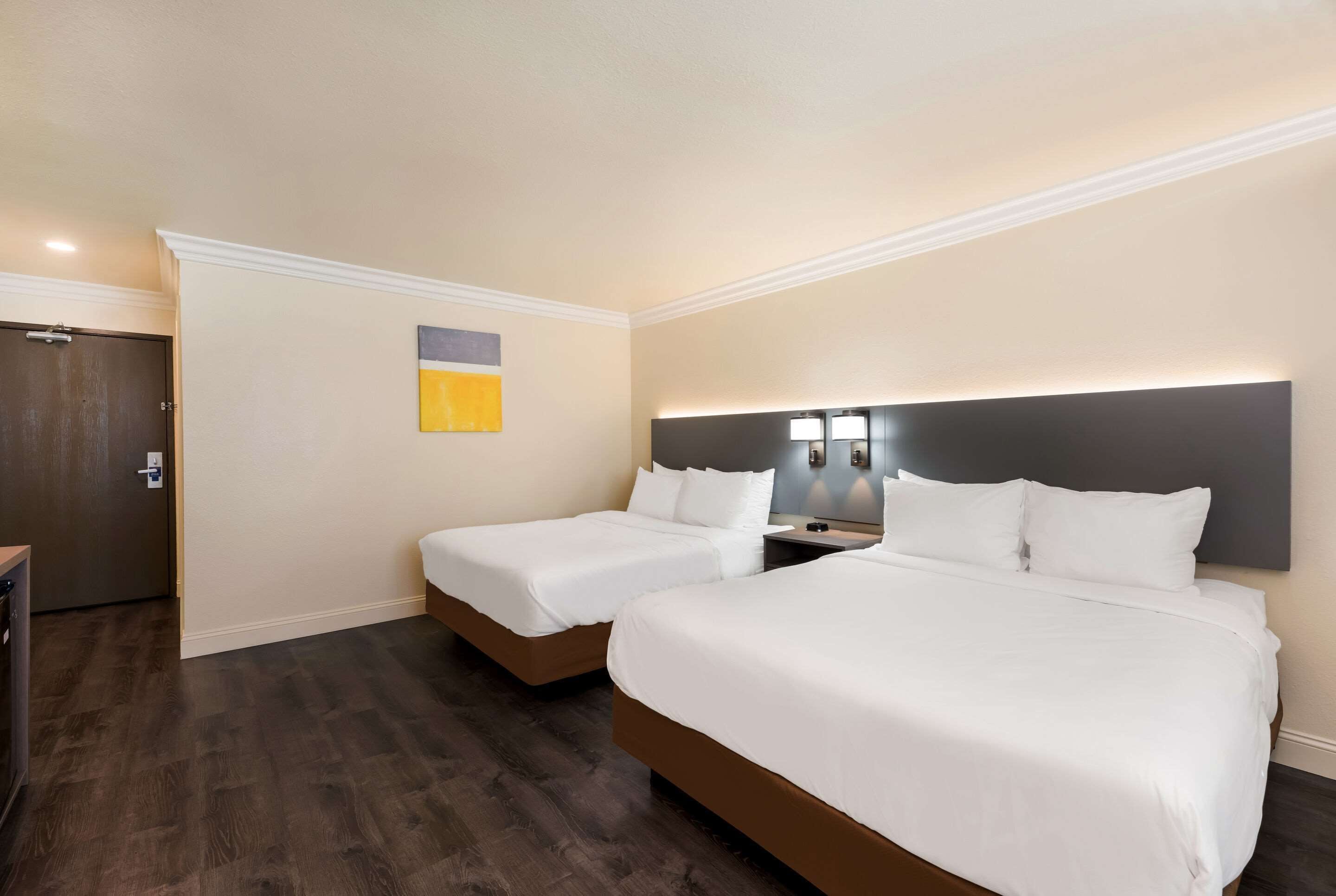 Une chambre d’hôtel avec deux lits, un parquet en bois foncé, une porte blanche et un petit tableau accroché au mur.