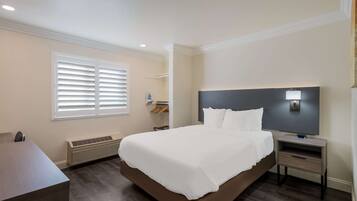 A hotel room with a large bed, a bedside table, a desk, a window with blinds, a wall-mounted TV, and a ceiling light.