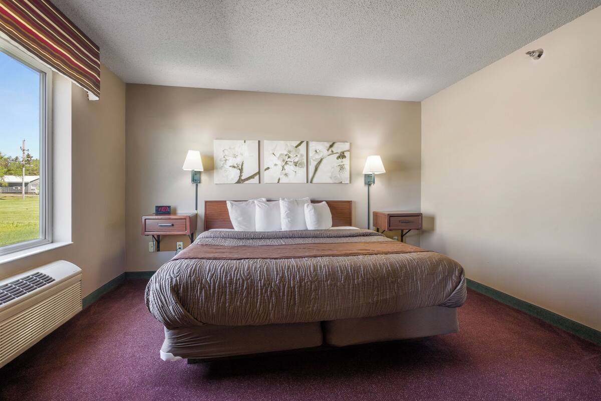 A hotel room with a large bed, two bedside tables, a window with a view, and a wall-mounted air conditioner.