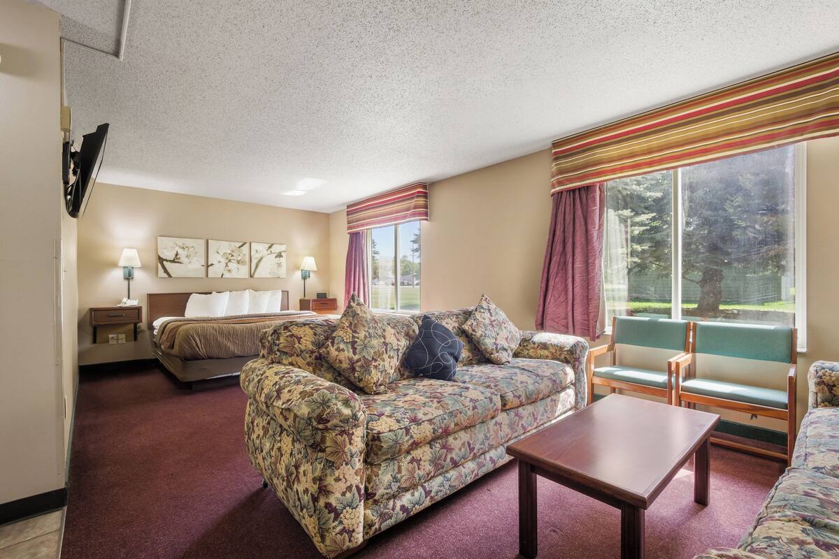 A hotel room with a floral sofa, a wooden coffee table, a bed with a headboard, a nightstand with a lamp, and a window with curtains.