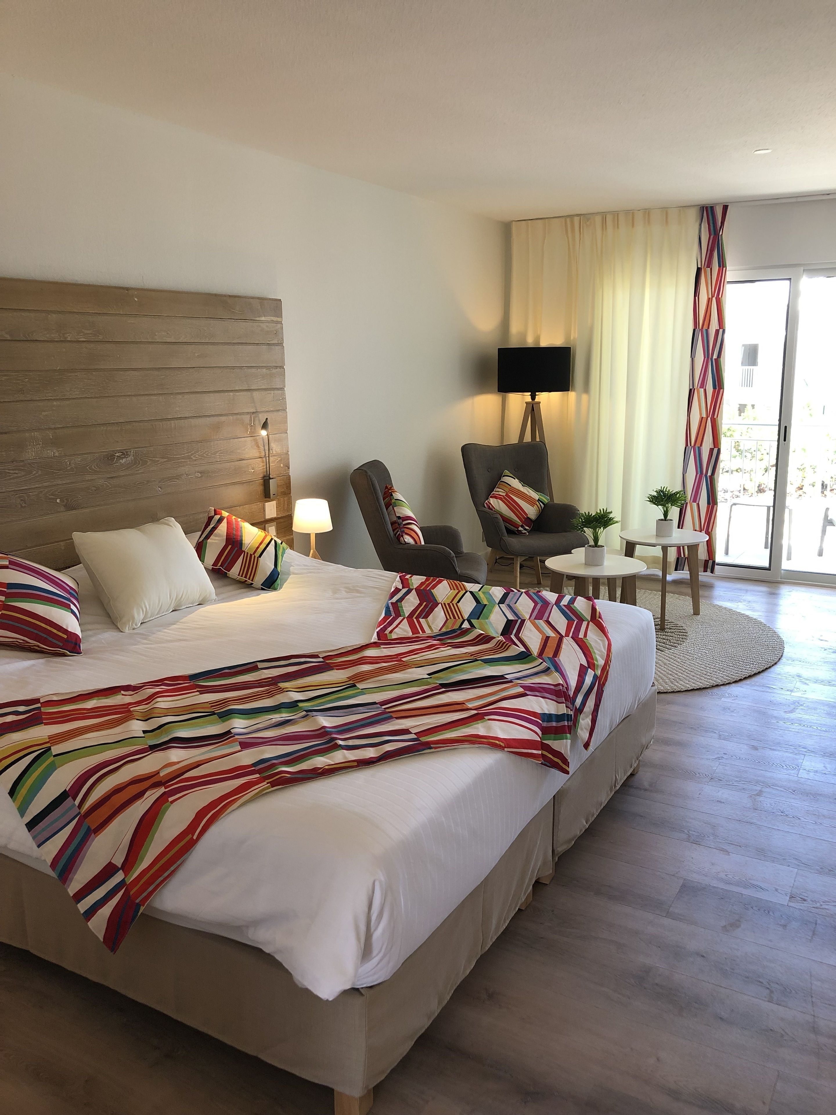 A hotel room with a large bed, two armchairs, a small table, a lamp, and a view of the outdoors.