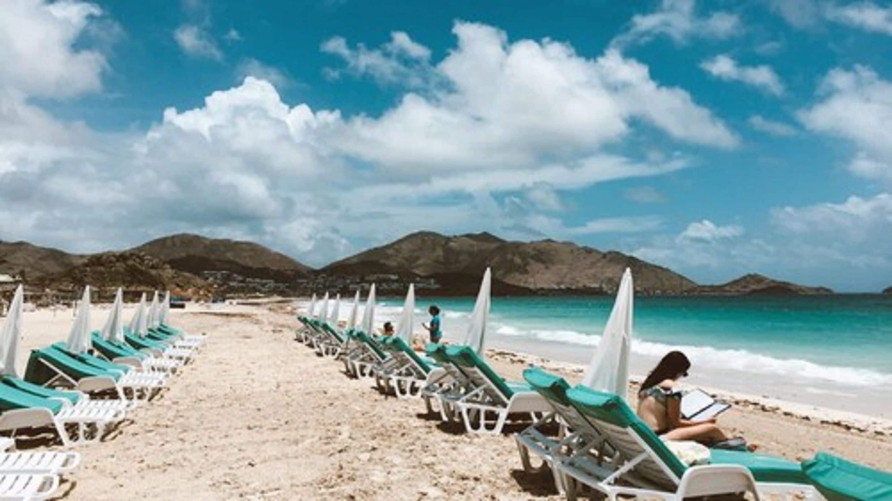 Uma praia com fileiras de espreguiçadeiras e guarda-sóis, céu claro e montanhas ao fundo.