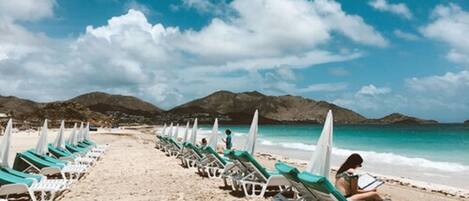A beach with rows of sun loungers and umbrellas, a clear sky, and mountains in the background.