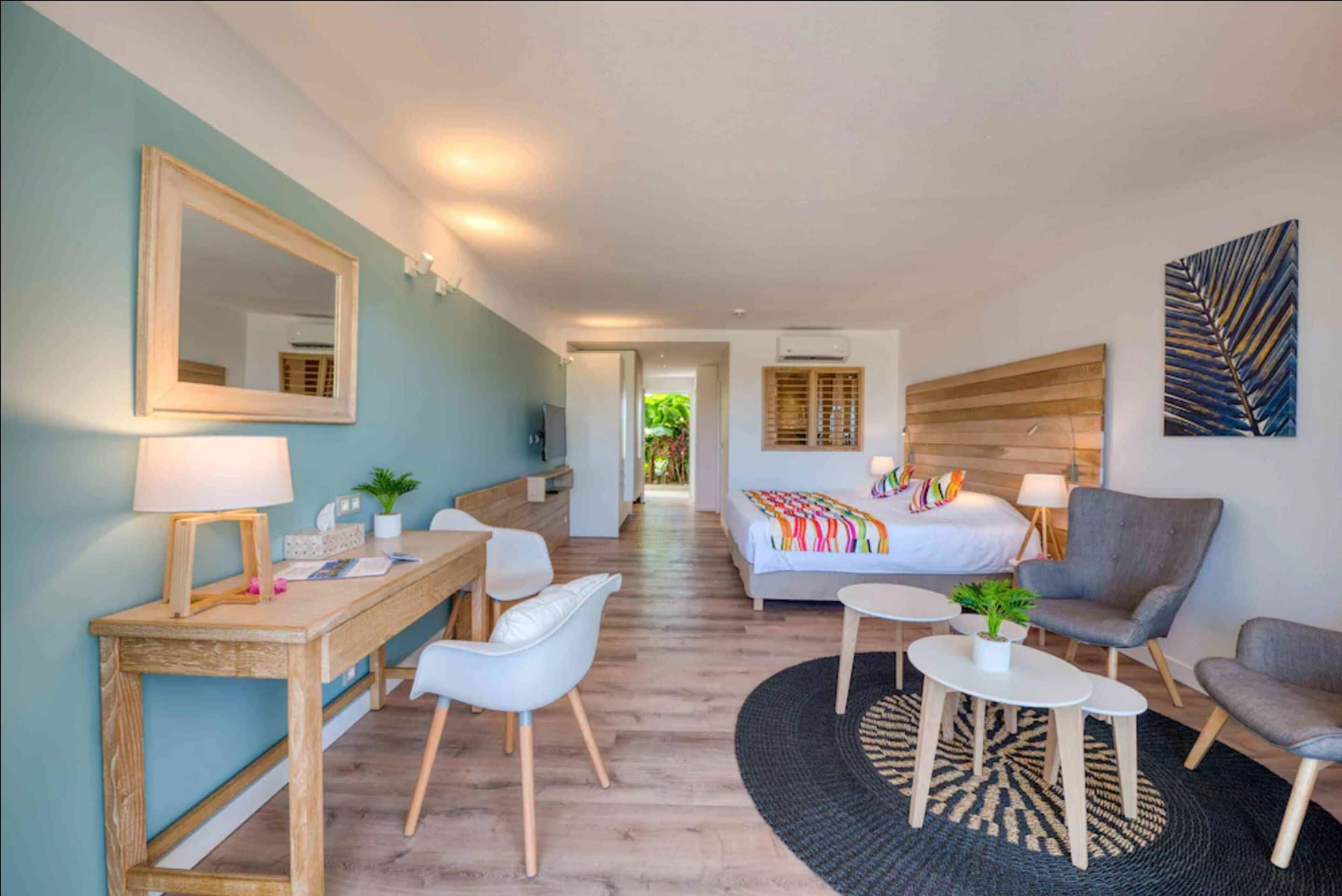 Un hotel moderno con un escritorio de madera, sillas blancas, una cama con ropa de cama colorida, un sillón gris y una mesa de centro redonda.