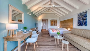 Una habitación de hotel moderna con un escritorio de madera, sillas blancas, una cama con manta colorida, un sofá color beige y un espejo grande en la pared.