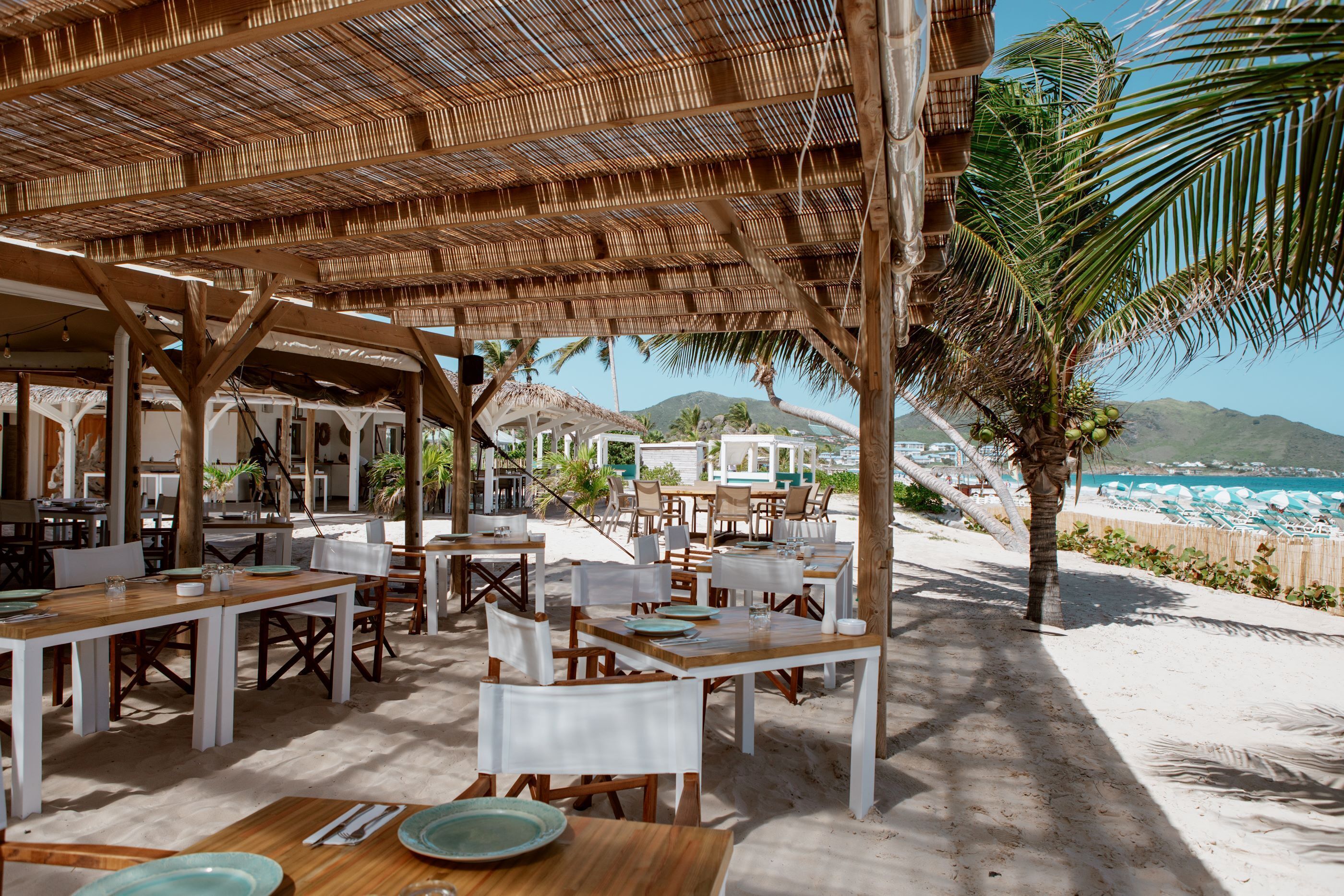 An outdoor dining area with wooden tables and white chairs, a thatched roof, and a view of the beach and mountains in the background.