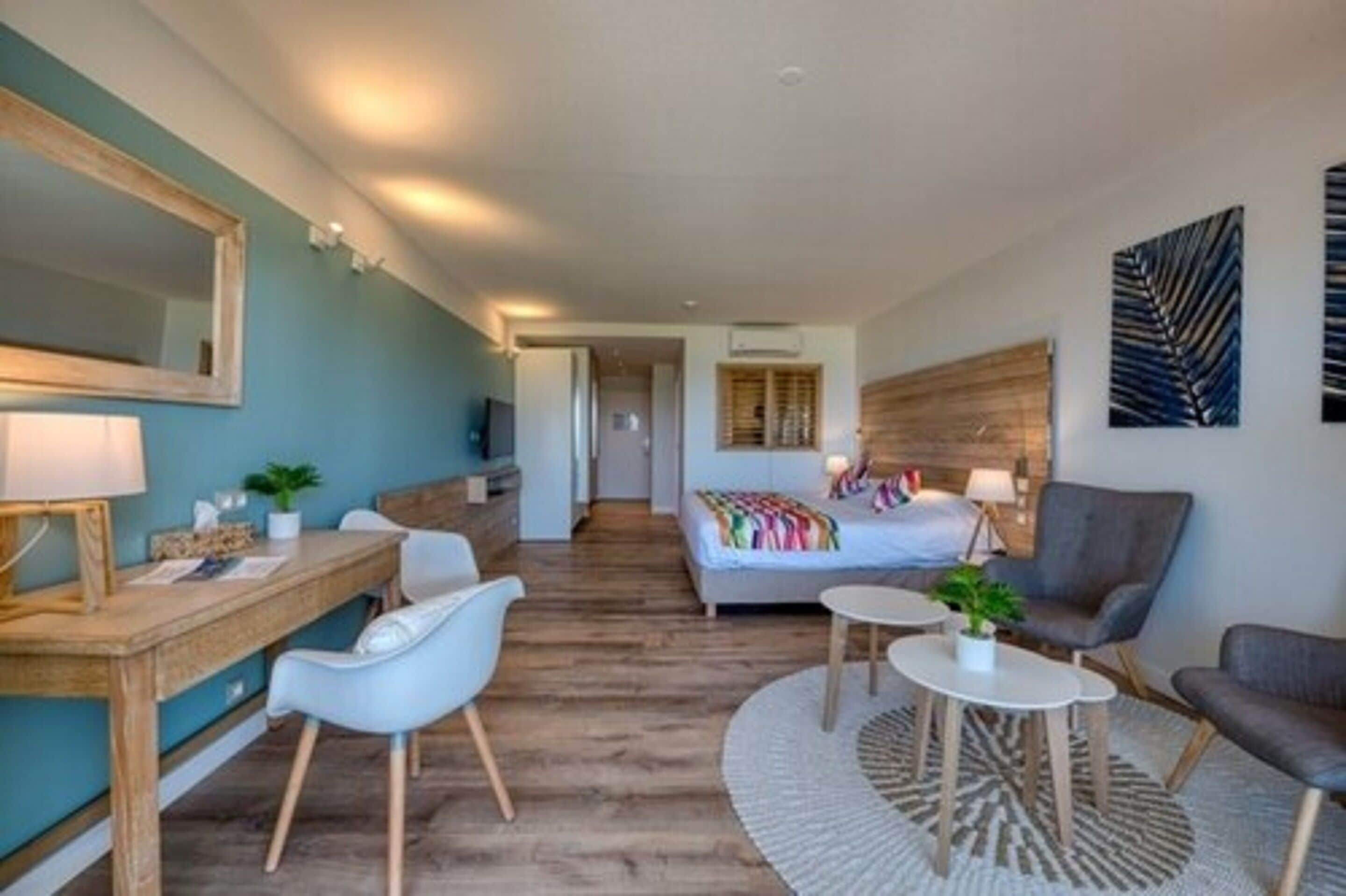 Ein modernes Hotelzimmer mit Schreibtisch aus Holz, einem Bett mit bunter Bettwäsche, einem großen Spiegel und einem Wandbild.