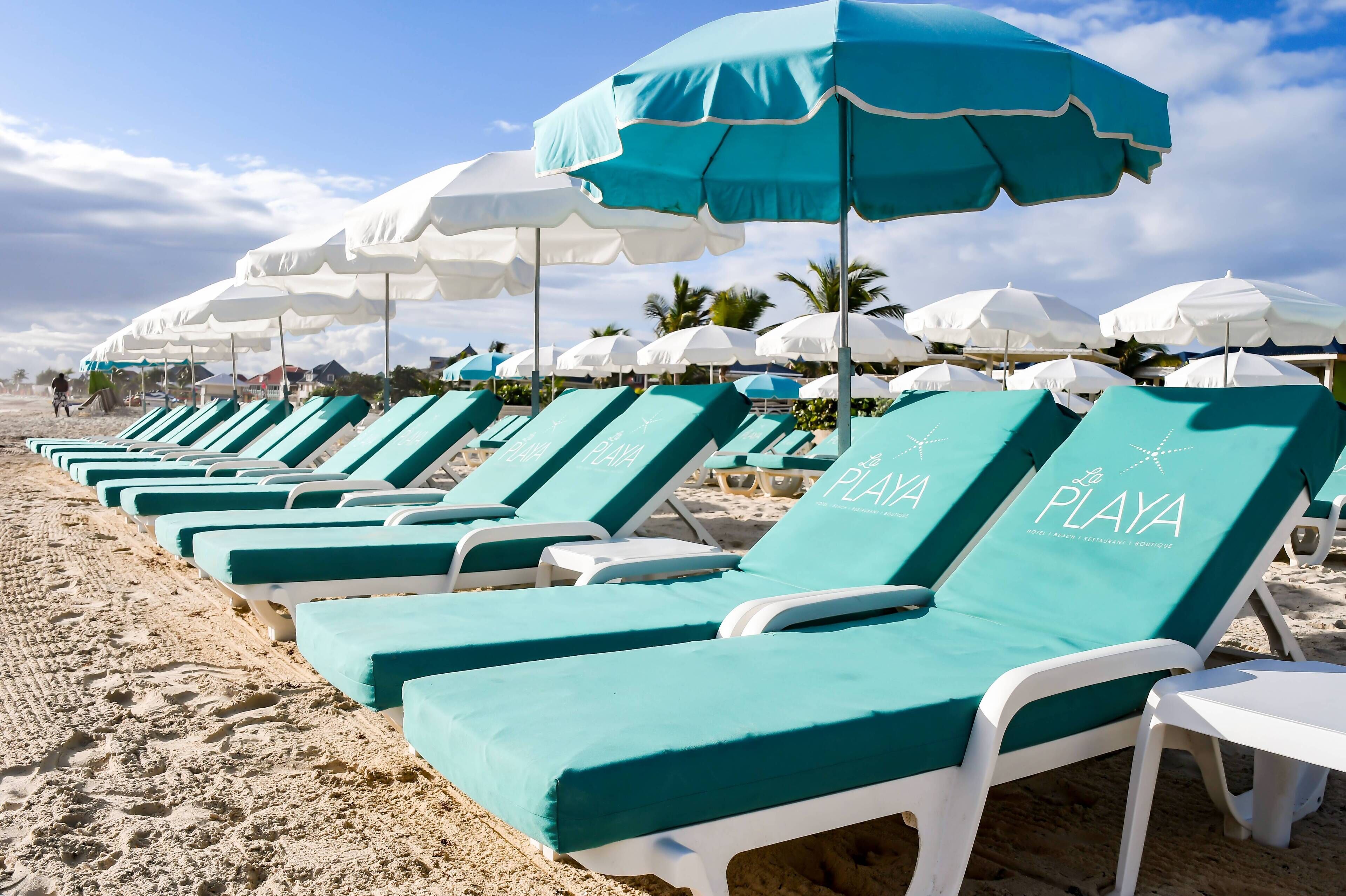 Una hilera de sillas de playa con la marca 'LA PLAYA', sombrillas blancas y una playa arenosa al fondo.