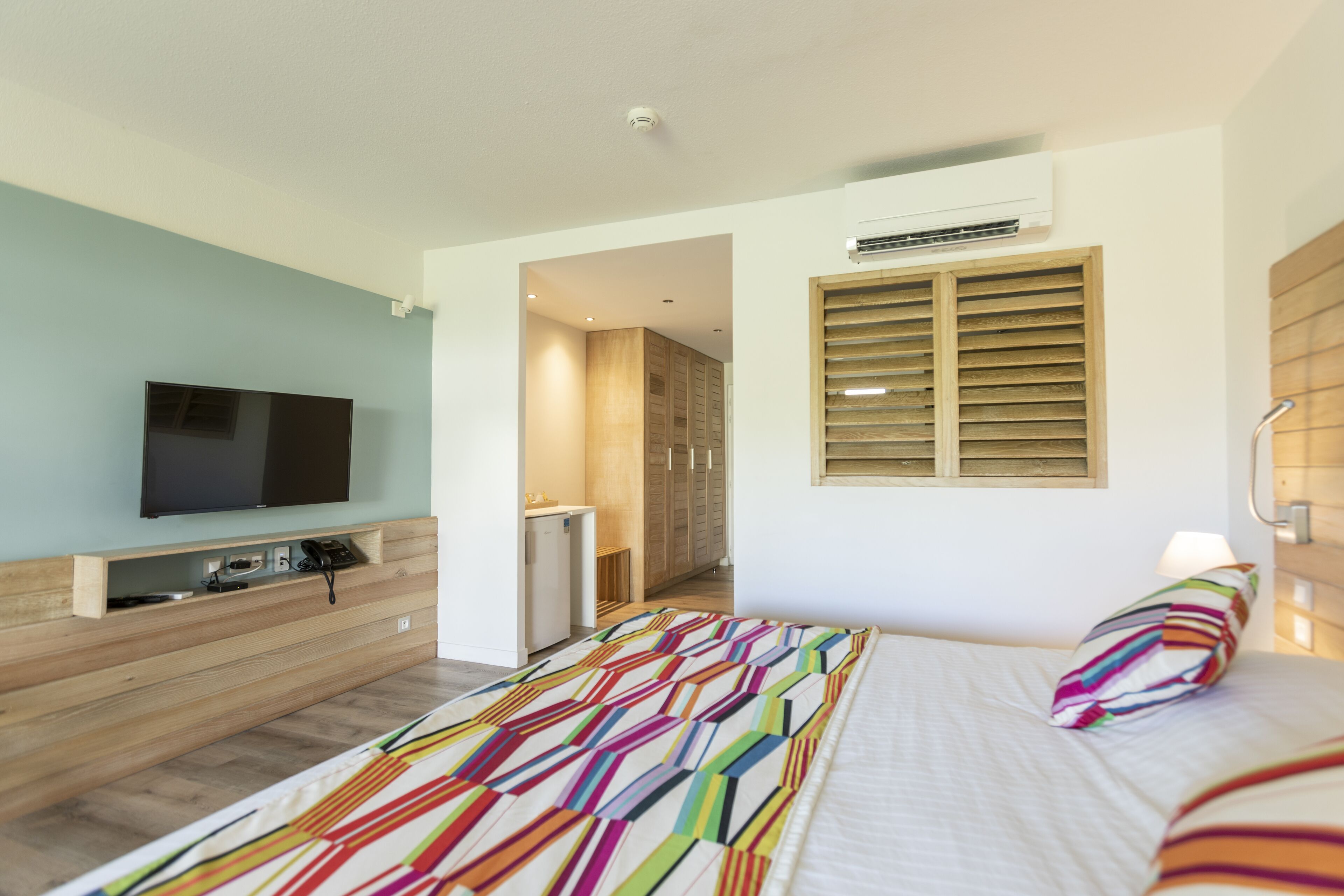 A modern hotel room with a large bed, a flat-screen TV mounted on a wooden console, a small kitchenette, and a bathroom visible through an open door.