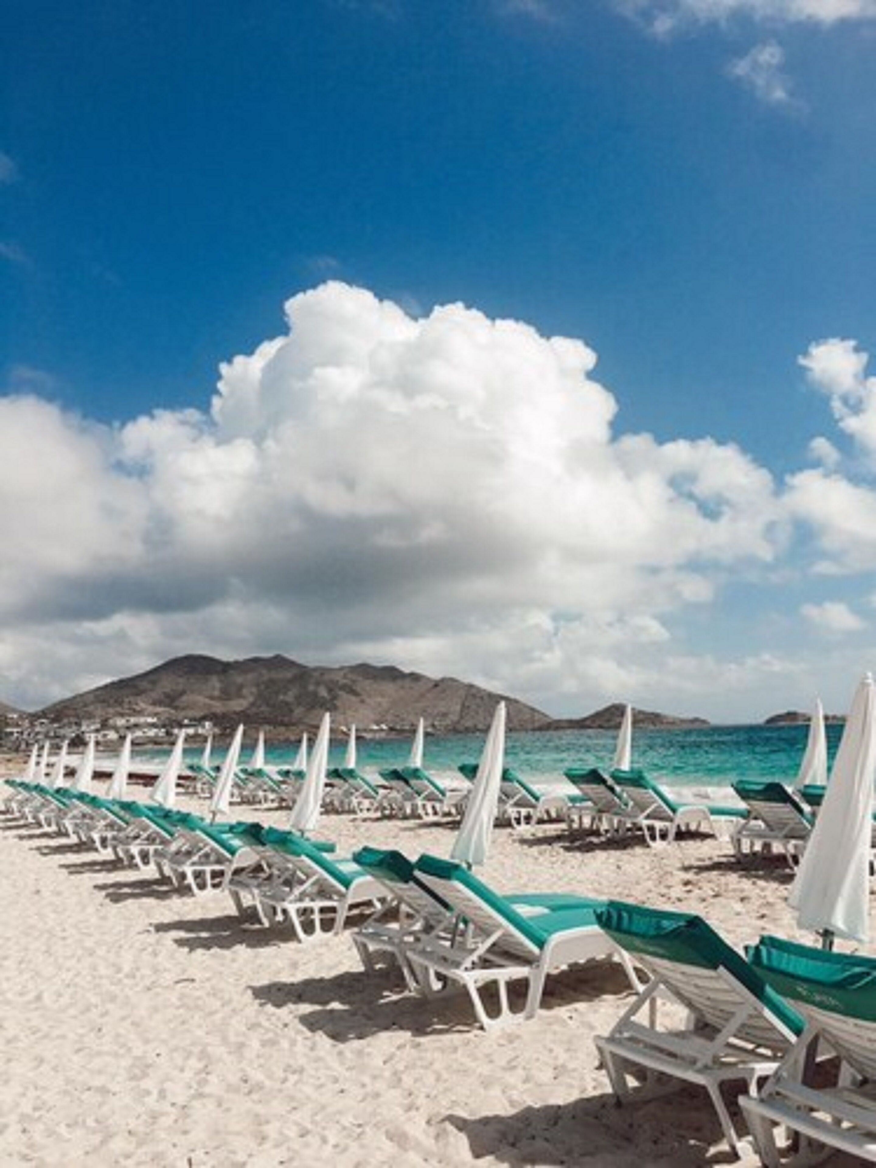 Ein Strand mit Reihen aus weißen Liegestühlen und grünen Handtüchern, einem klaren, blauen Himmel und Bergen im Hintergrund.