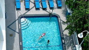 Ein Schwimmbecken mit einer schwimmenden Person und einer weiteren im Wasser, umgeben von blauen Liegestühlen und einem Baum.
