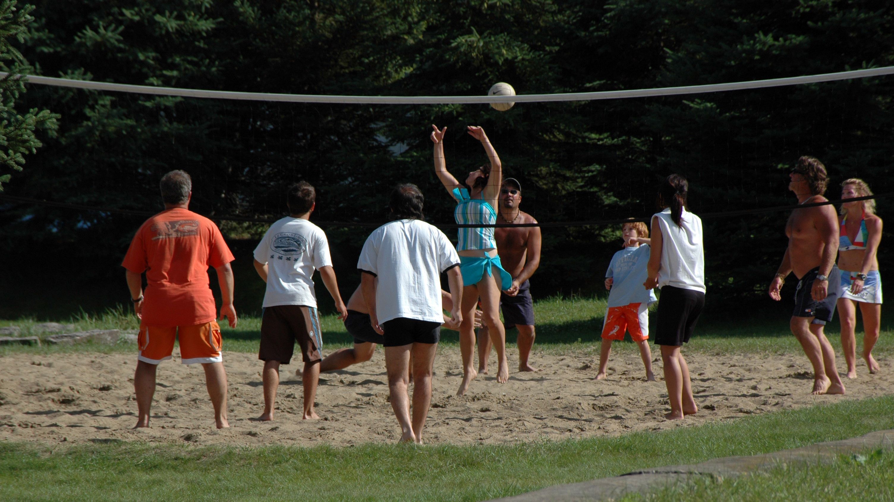 A group of people playing beach volleyball on a sandy court with trees in the background.