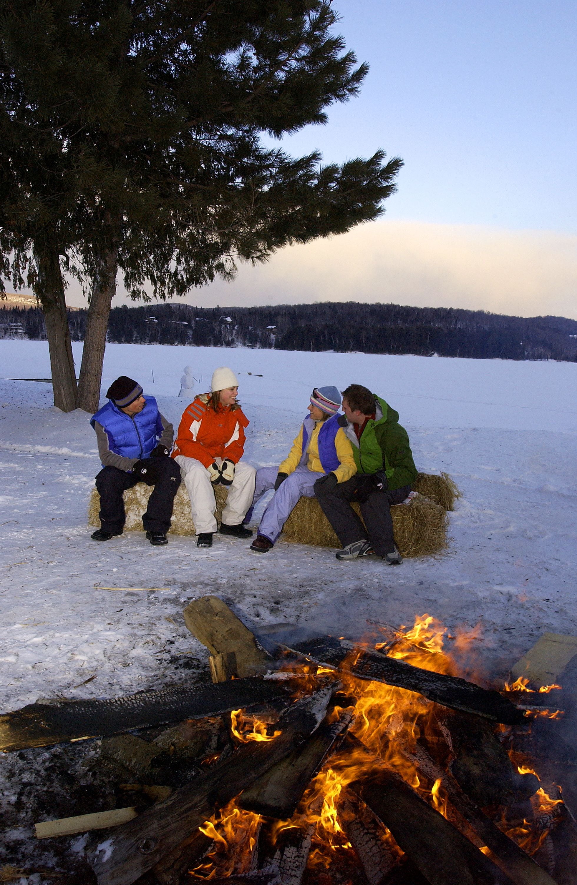 Three people sitting on a hay bale in a snowy outdoor setting with a fire in the foreground.