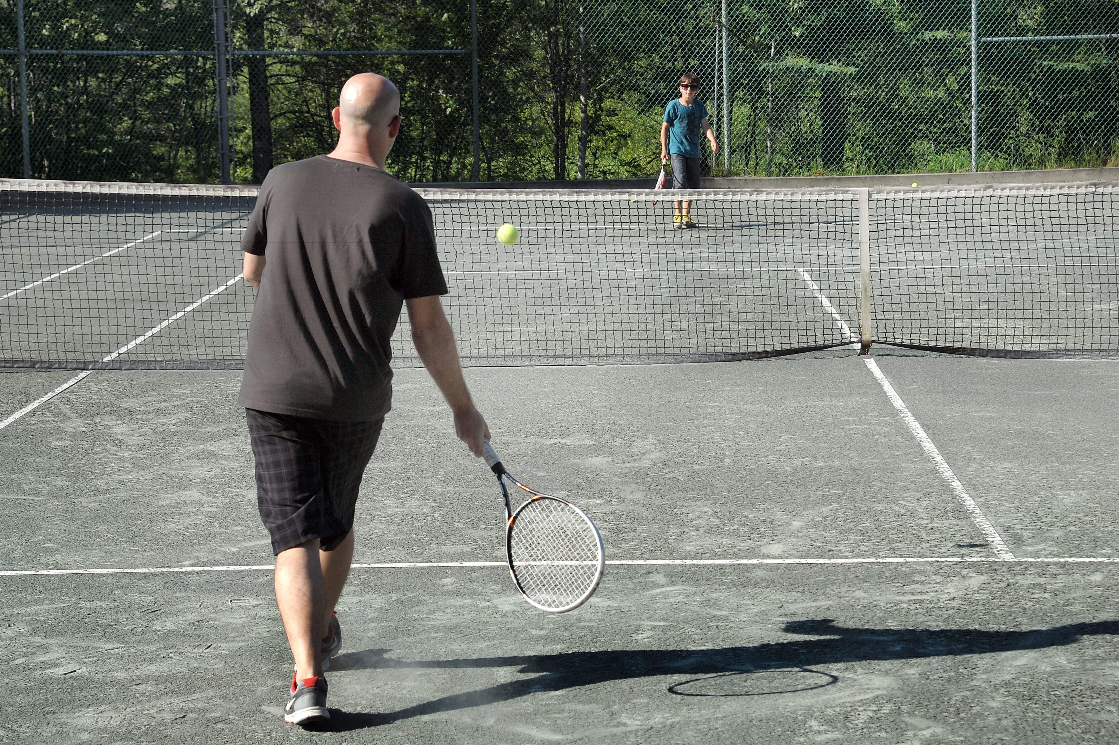 Ein Tennisplatz mit Netz, ein Spieler in schwarzem T-Shirt und Shorts, sowie ein weiterer Spieler im Hintergrund.
