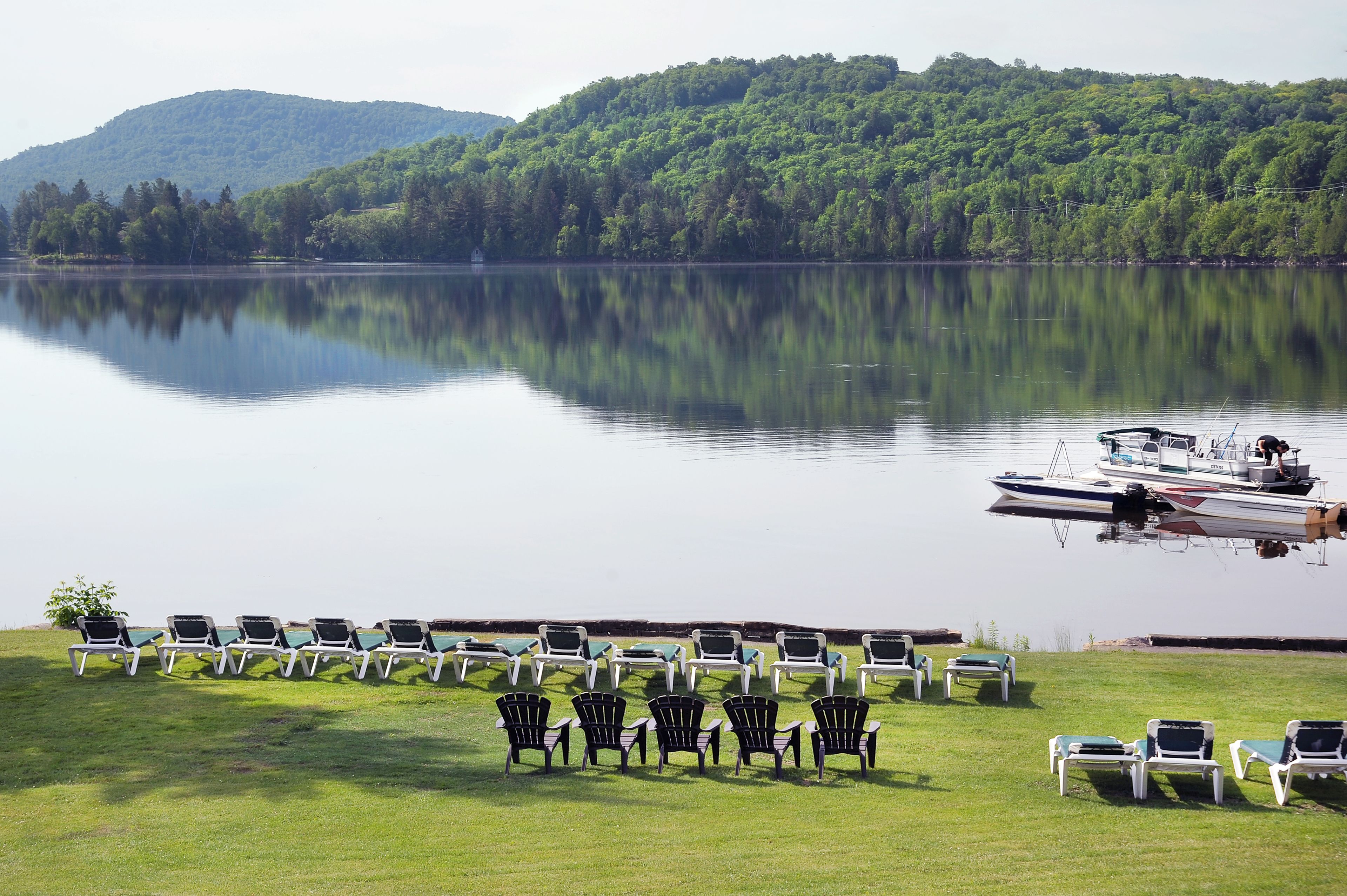 A lakeside view with a boat docked near the shore, surrounded by rows of lawn chairs and a grassy area.