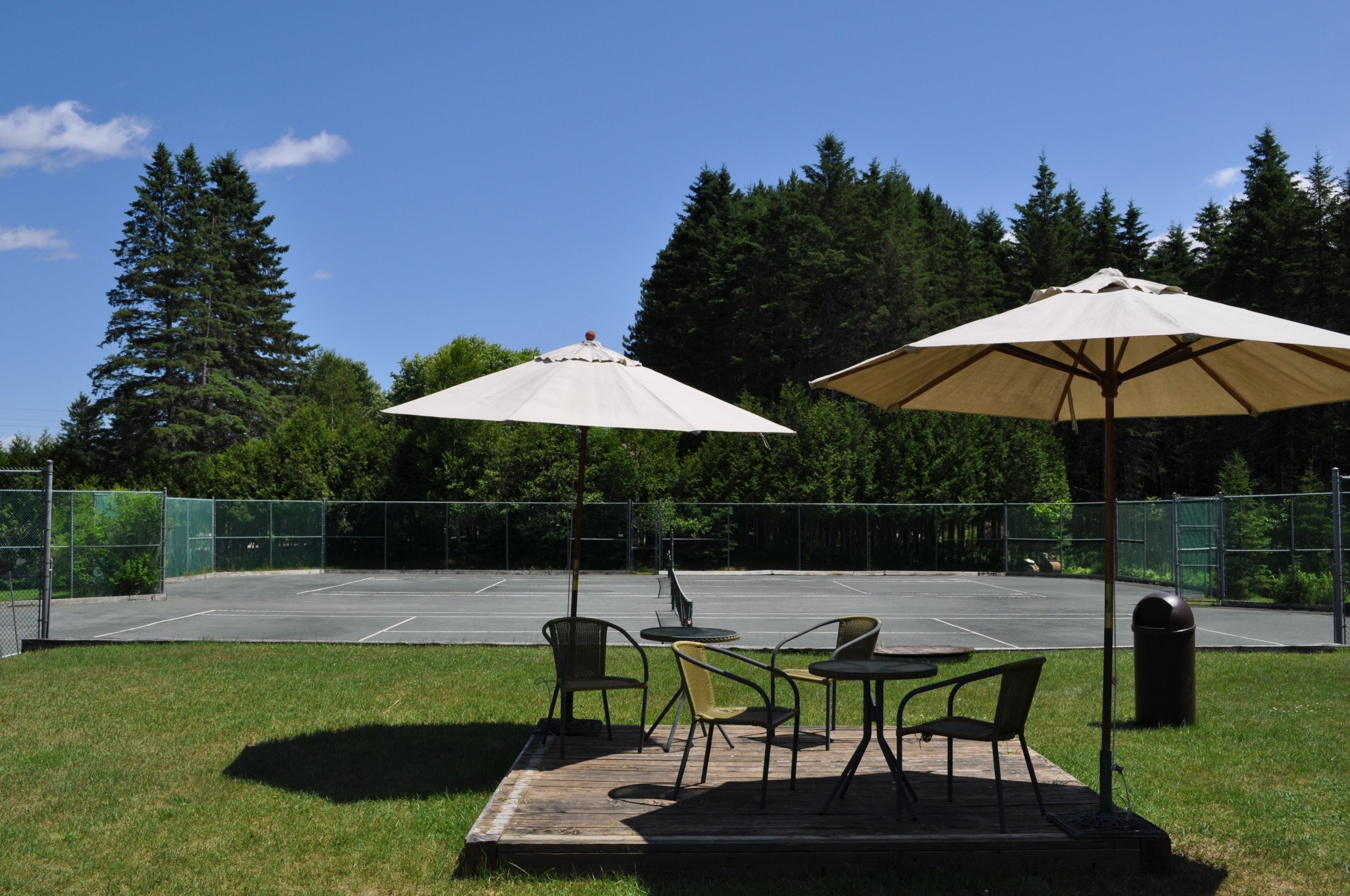 Un espace extérieur aménagé avec des tables et des chaises sous des parasols, un court de tennis et un paysage boisé en arrière-plan.