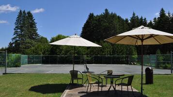 Un espace extérieur aménagé avec des tables et des chaises sous des parasols, un court de tennis et un paysage boisé en arrière-plan.