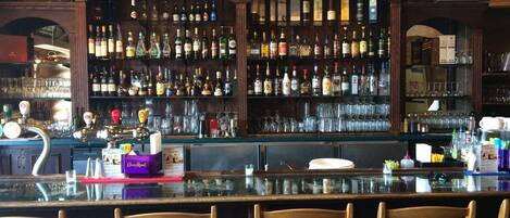 A bar with wooden shelves stocked with various liquor bottles, a bar counter with stools, and a television mounted on the wall.